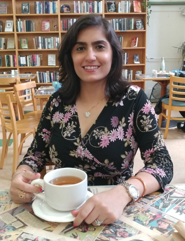 An image of the blog author sitting inside with a cup of tea with bookshelves and tables behind her.