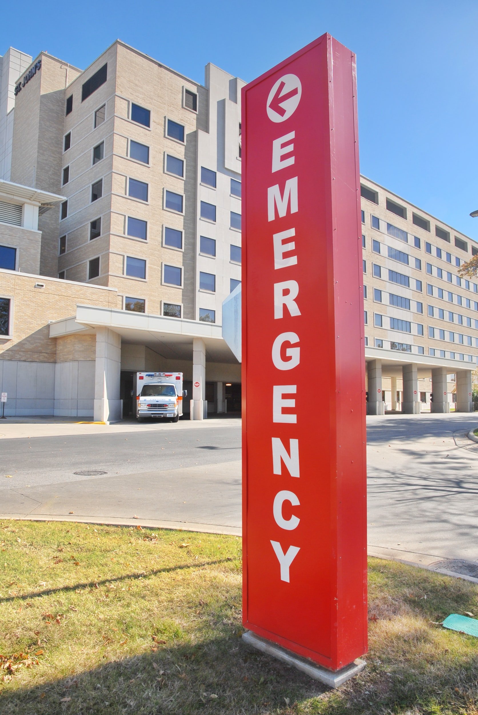 A red signs reads ‘emergency’ in white writing. There is a building and an ambulance behind the sign.