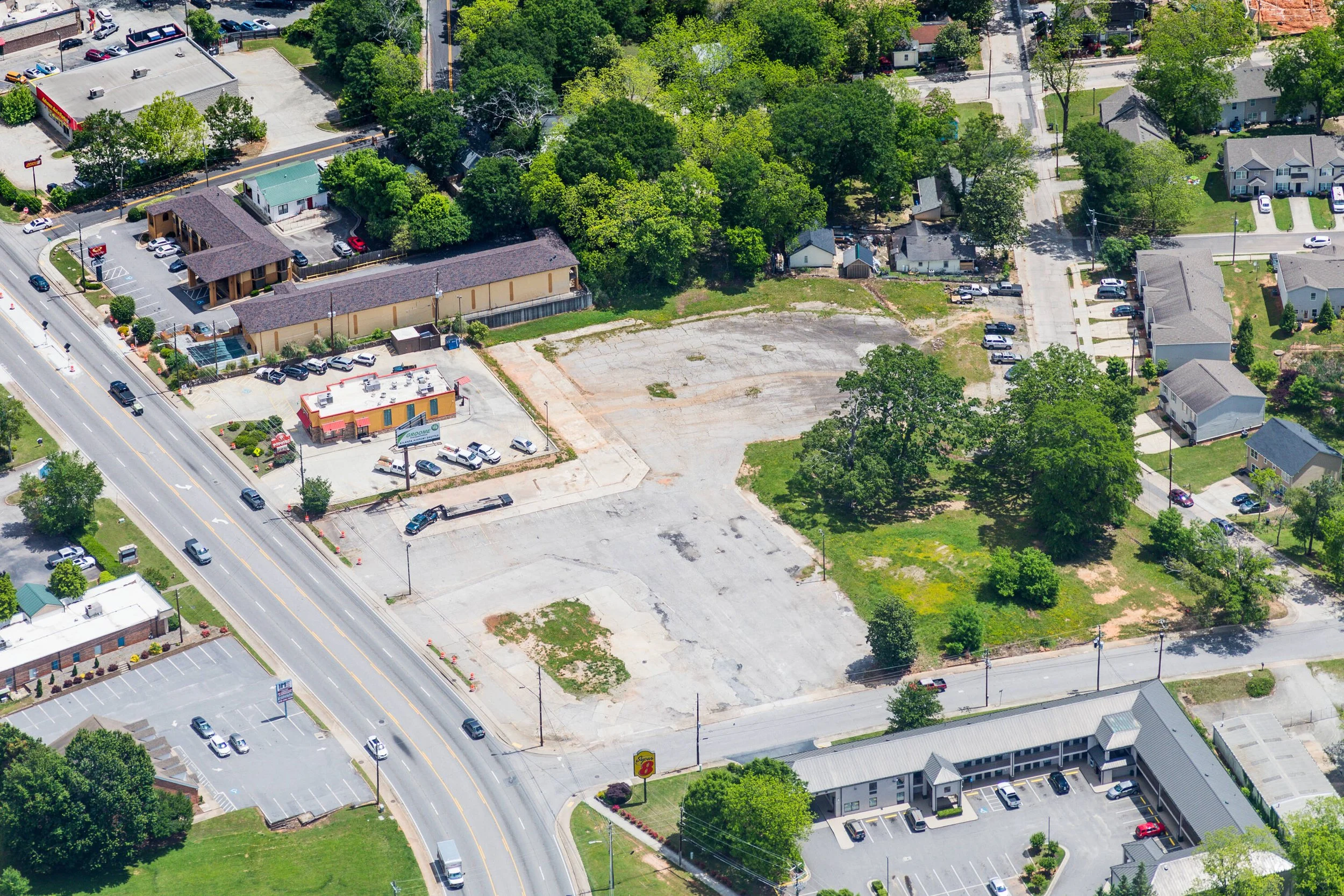 An aerial view of a commercial area with a vacant lot, surrounded by parking lots, small buildings, houses, and trees.