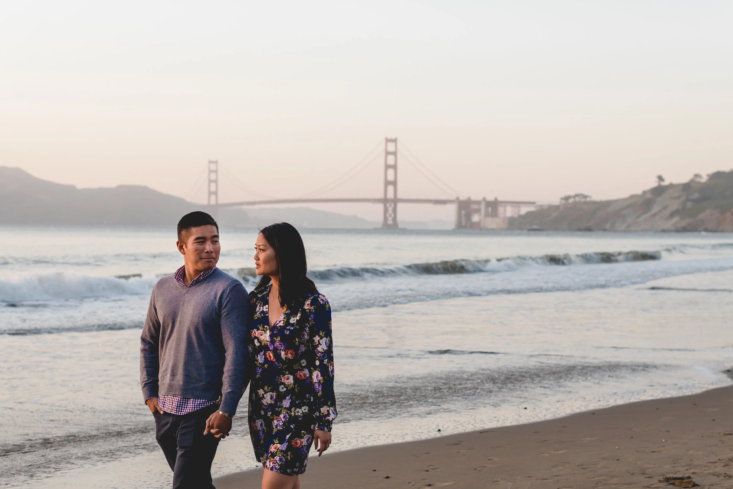 A young couple walking hand in hand along the beach at sunset with the Golden Gate Bridge visible in the background.