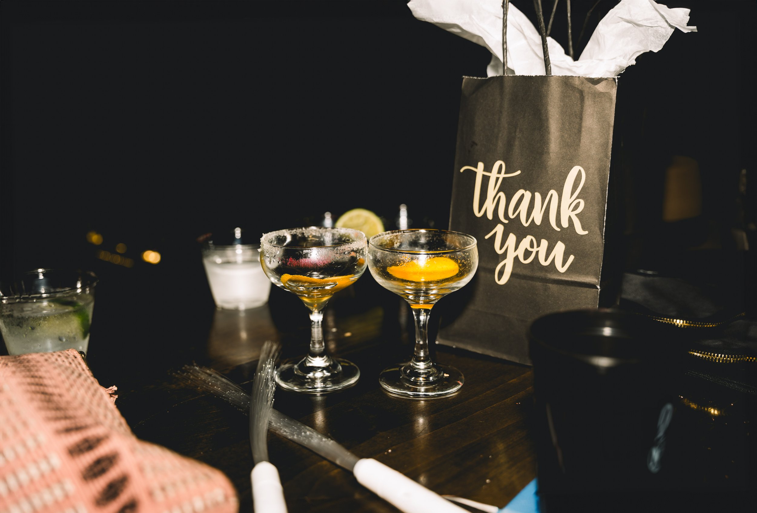Two glasses with lemon slices on a dark table, behind a black paper bag with 'thank you' written on it, surrounded by colorful items and a black background.