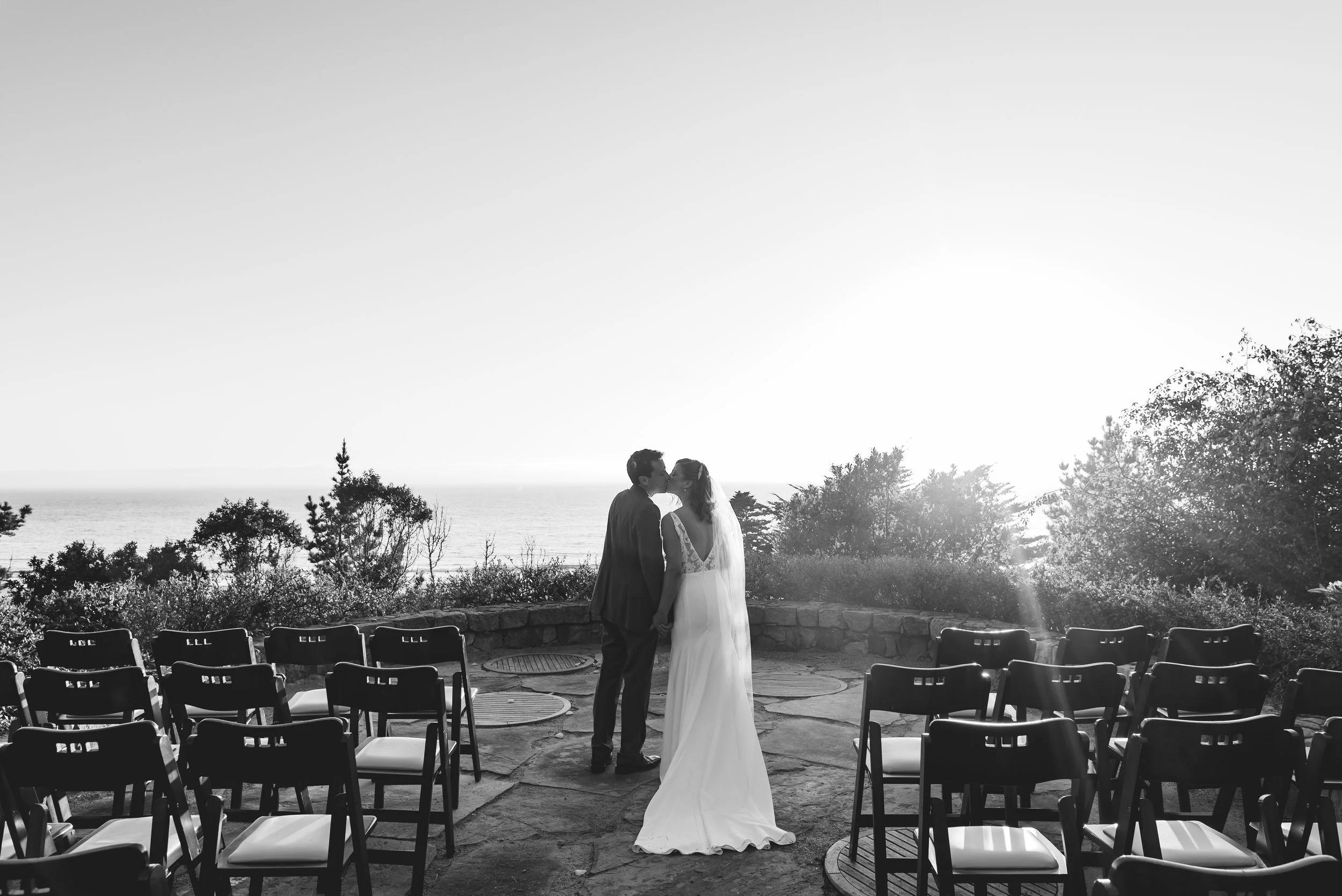 A bride and groom kissing during their wedding ceremony outdoors near the coast with empty chairs around them, with the ocean and trees in the background, in black and white.