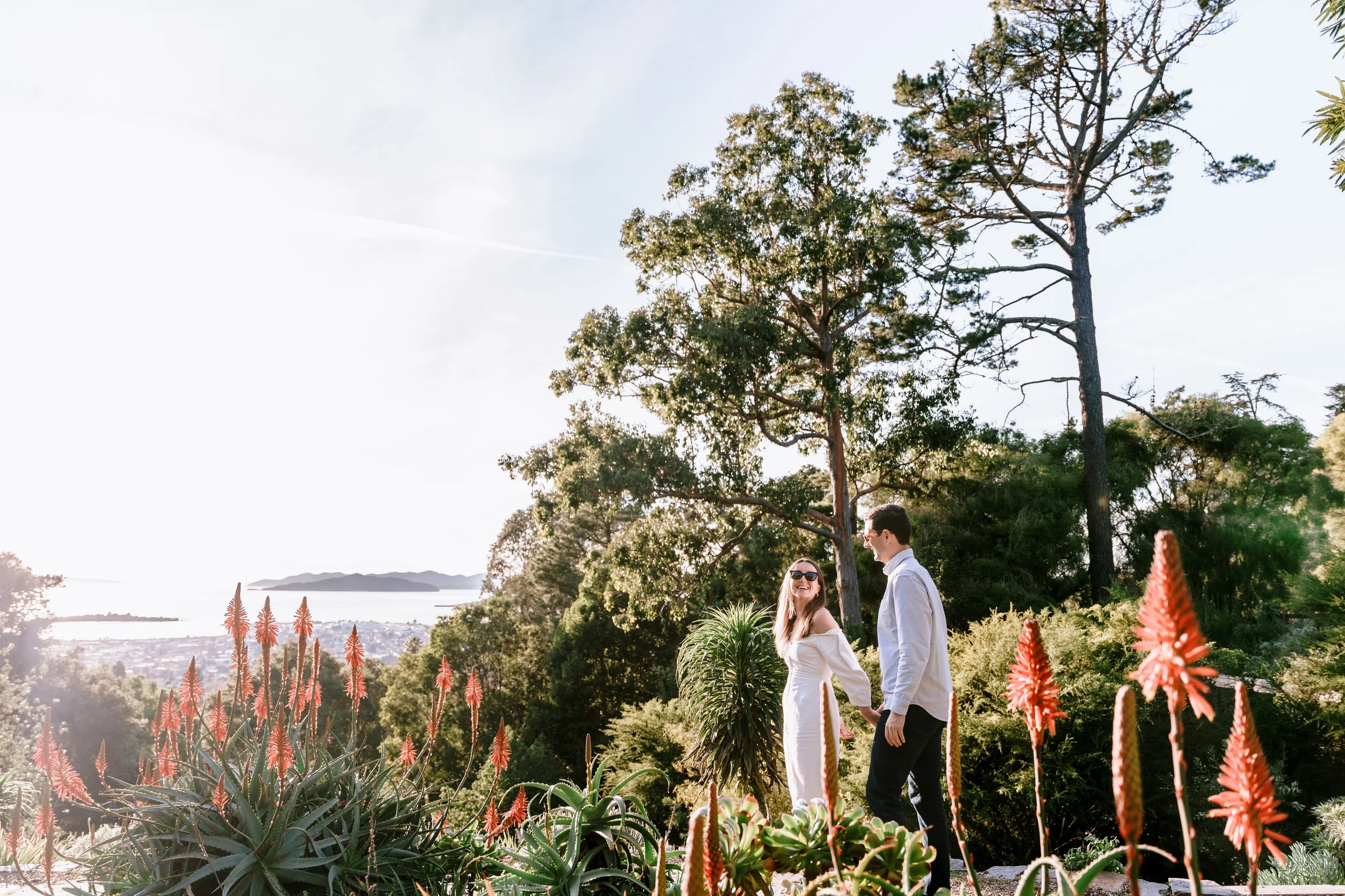 A couple holding hands and smiling in a garden with orange flowers, tall trees, and a view of water and distant islands in the background.
