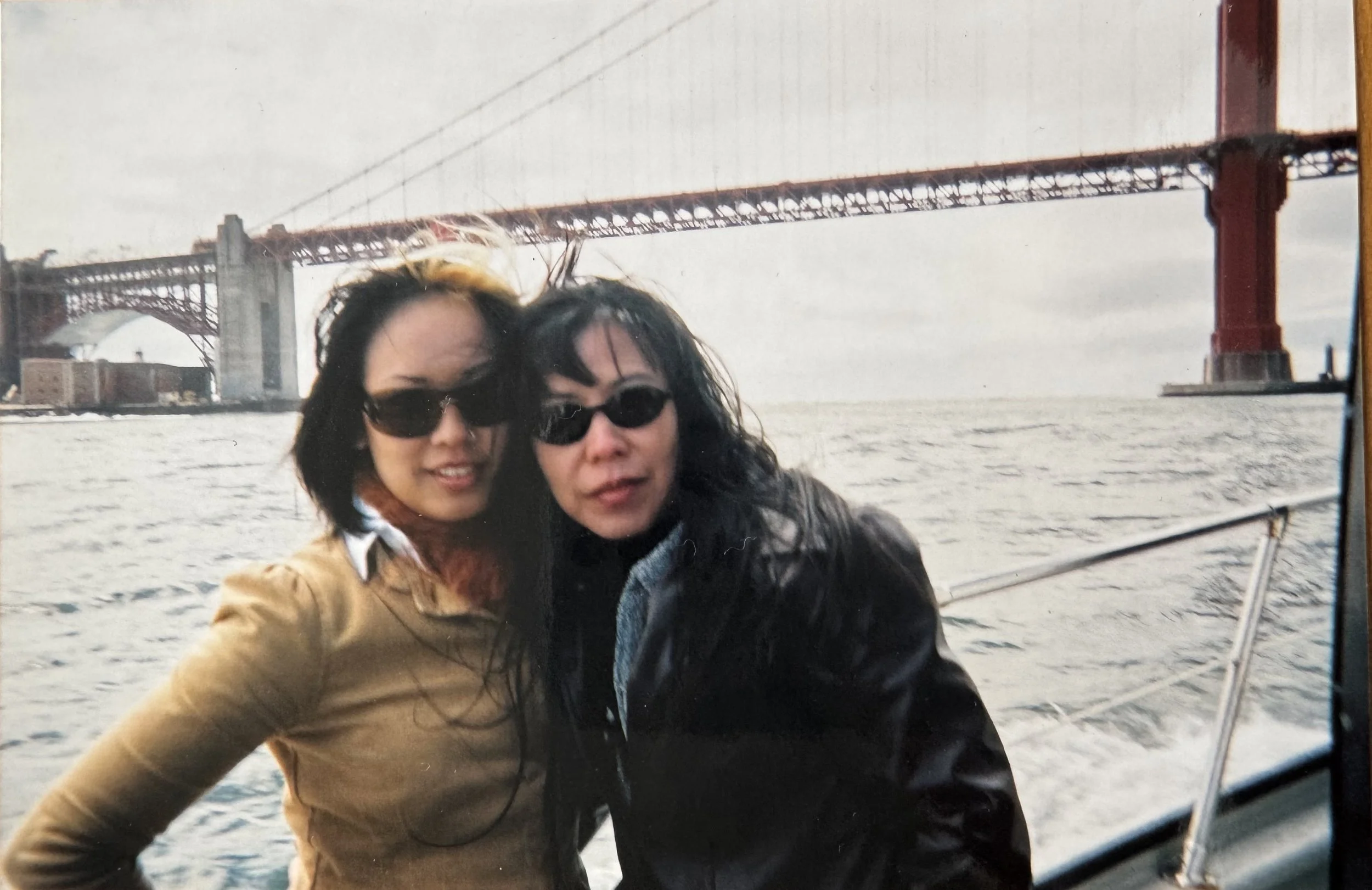 Two women with sunglasses posing on a boat with the San Francisco Bay Bridge in the background.