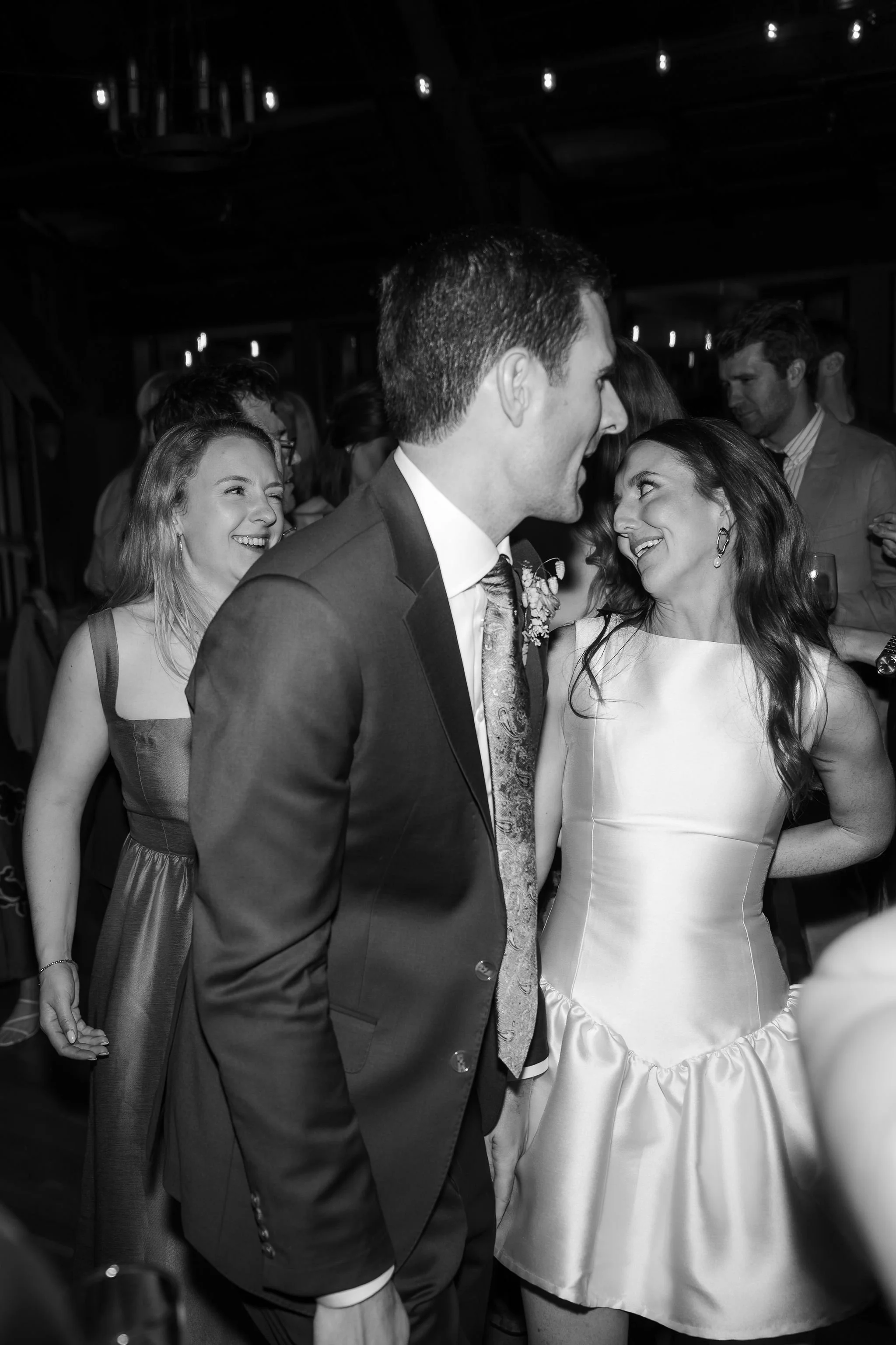Black and white photo of a wedding reception with a bride and groom looking at each other, surrounded by happy guests.
