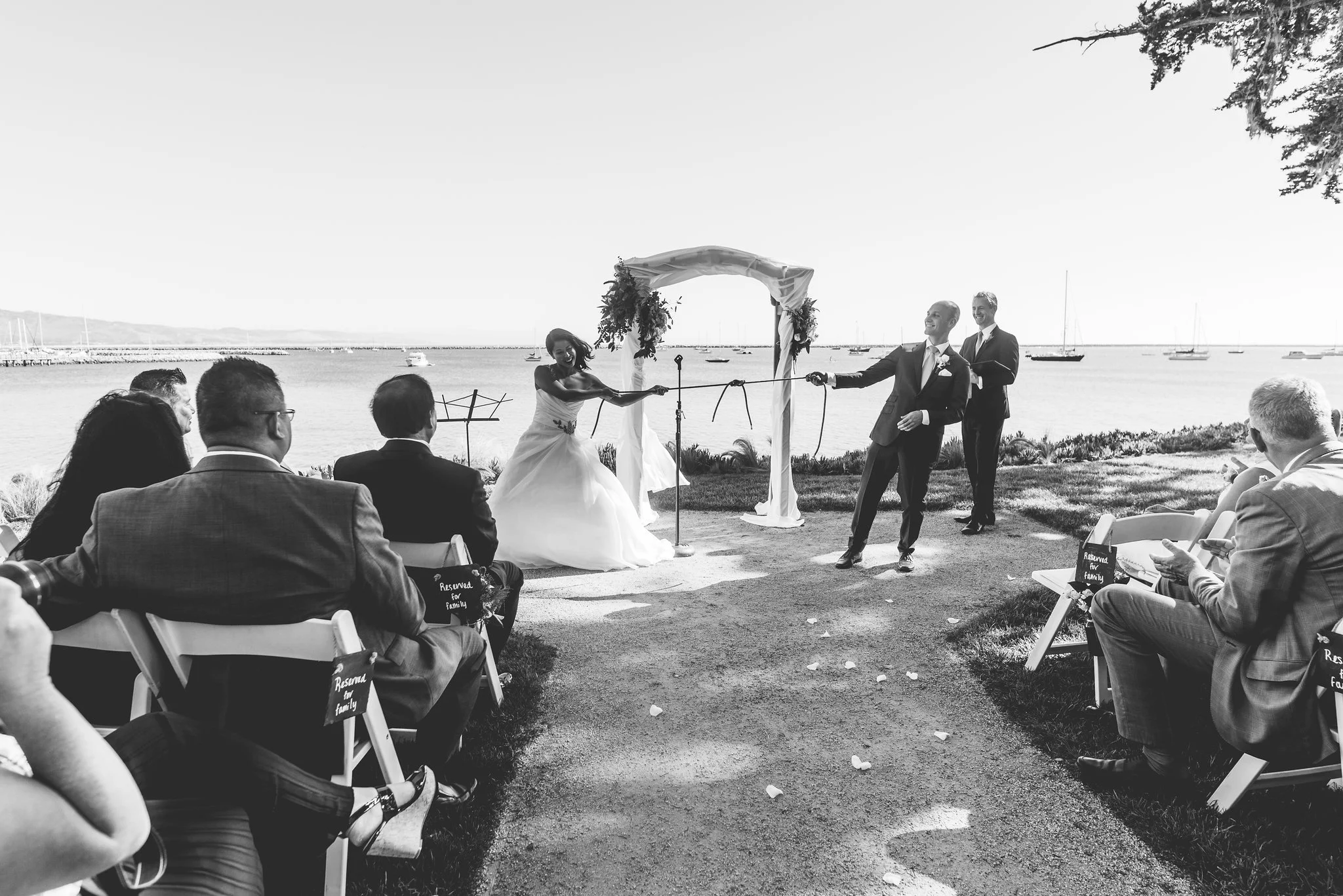 Black and white photo of a wedding ceremony outdoors near a body of water, where the bride and groom are pulling on a rope during their vows, with guests seated on either side and sailboats in the background.