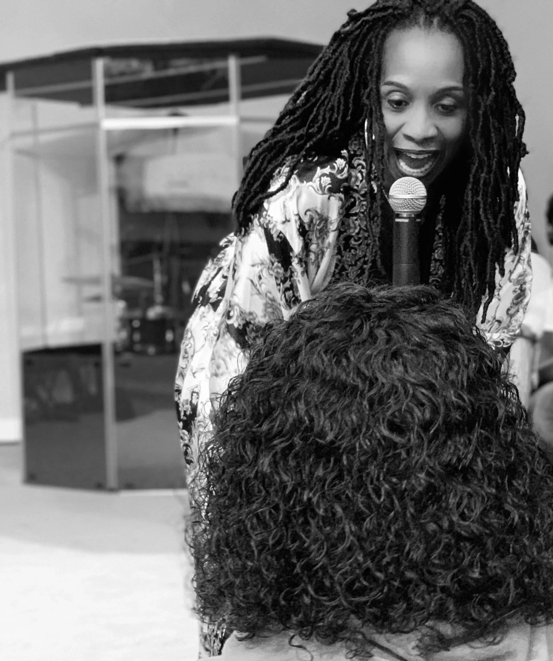 A black-and-white photograph of a woman with long, curly hair speaking into a microphone, appearing to be at a gathering or event.
