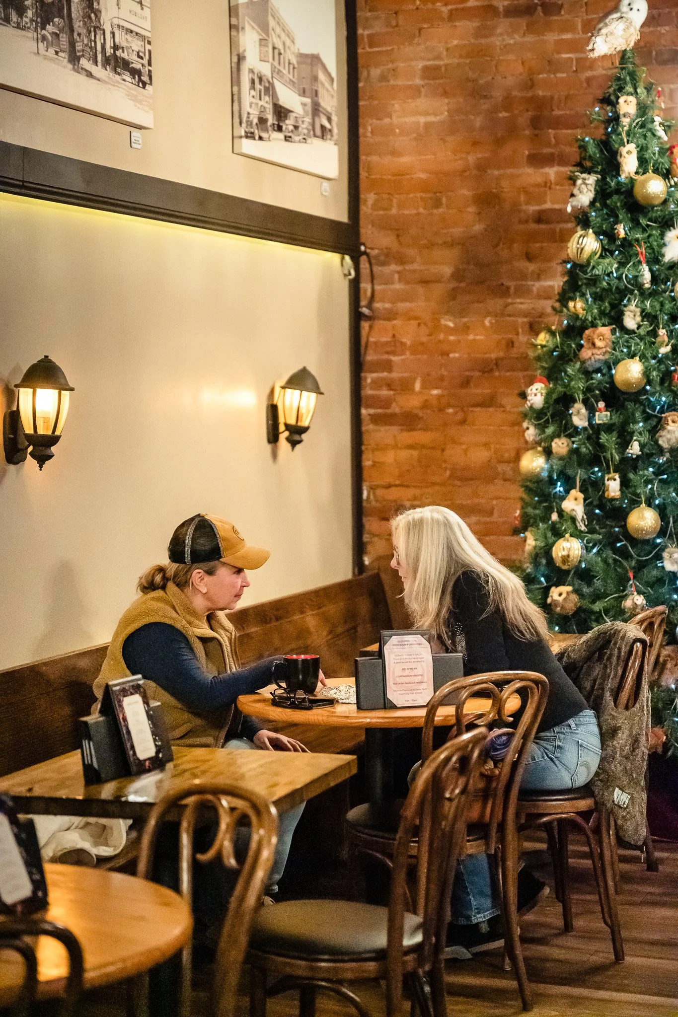Two women sitting at a small round table in a cozy restaurant with a decorated Christmas tree nearby, brick and beige walls, and warm lighting.