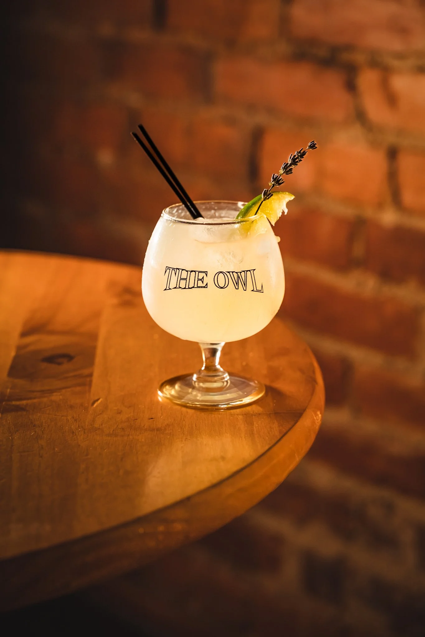 A cocktail in a stemmed glass with the words 'THE OWL' printed on it. The drink is garnished with a lemon wedge and a sprig of lavender, with two black straws. The glass is placed on a wooden surface with a brick wall background.