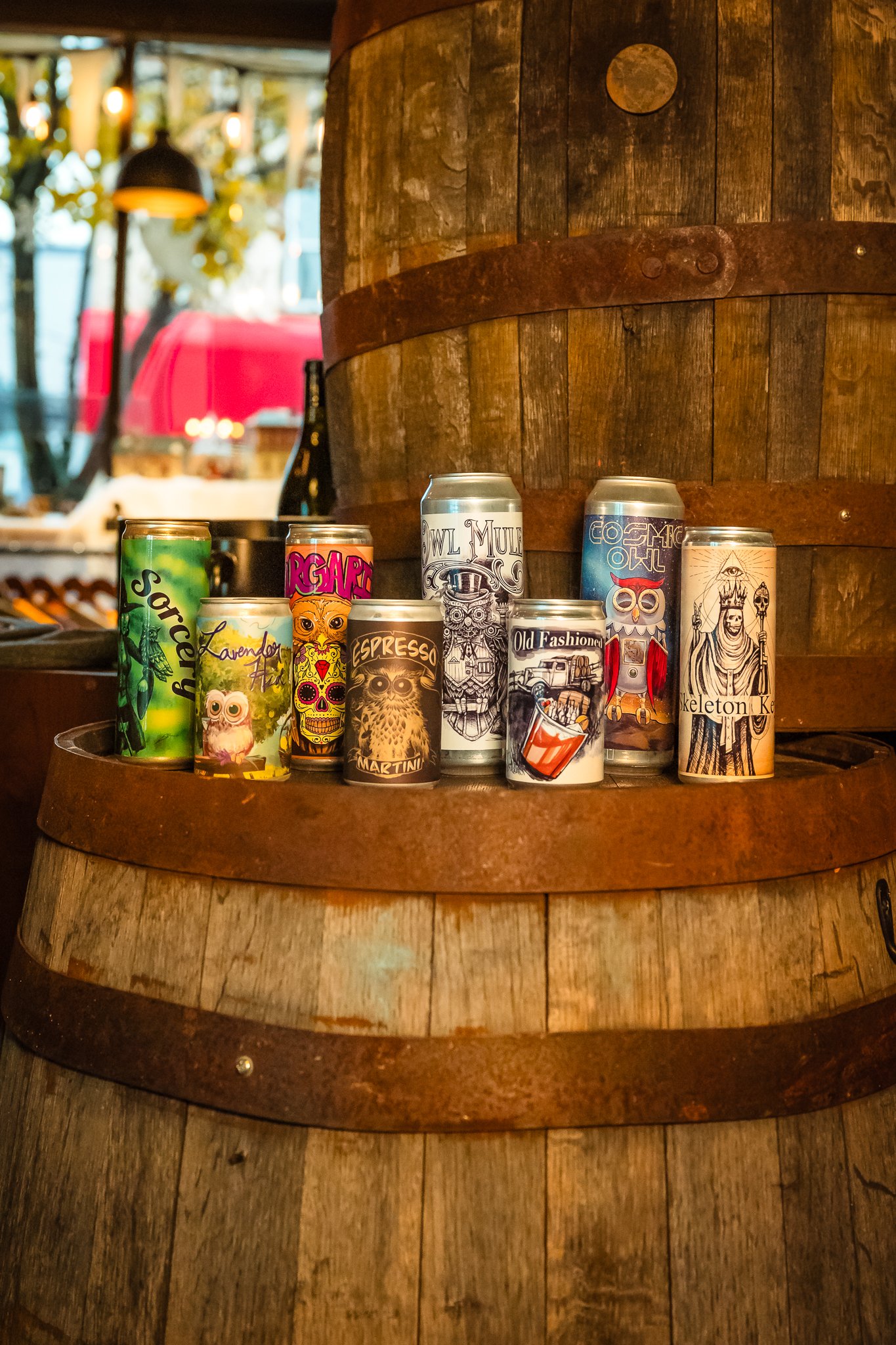 A collection of colorful cans with various artistic labels is displayed on a wooden barrel in a rustic setting.
