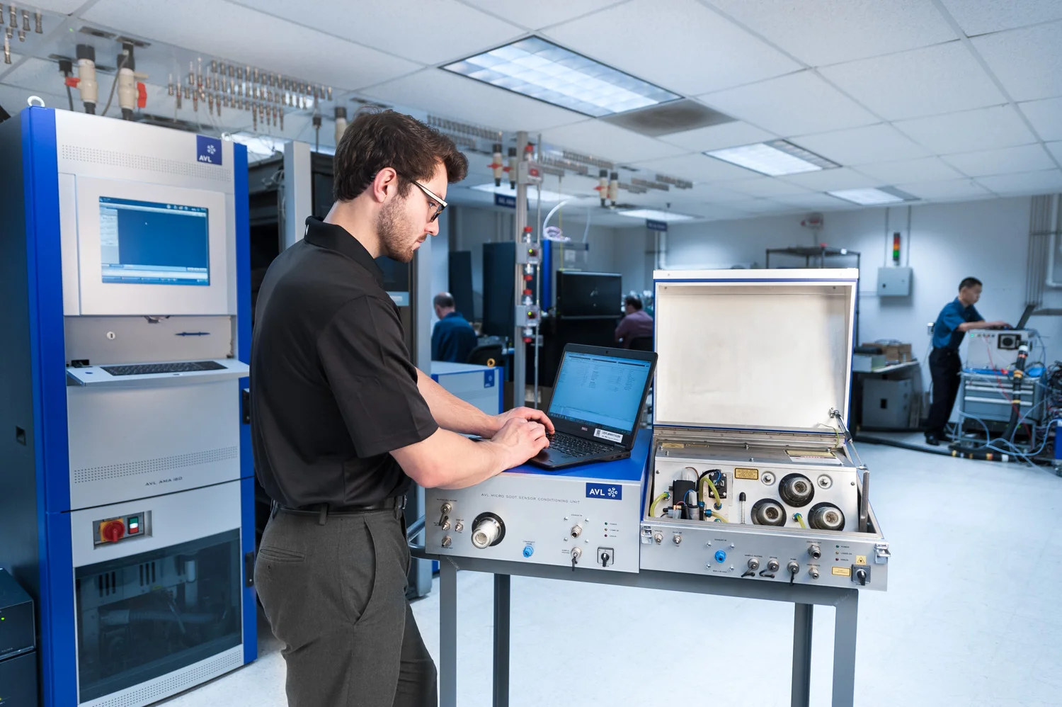 A man working on a laptop in a laboratory or technical workspace with electronic equipment and other people working in the background.