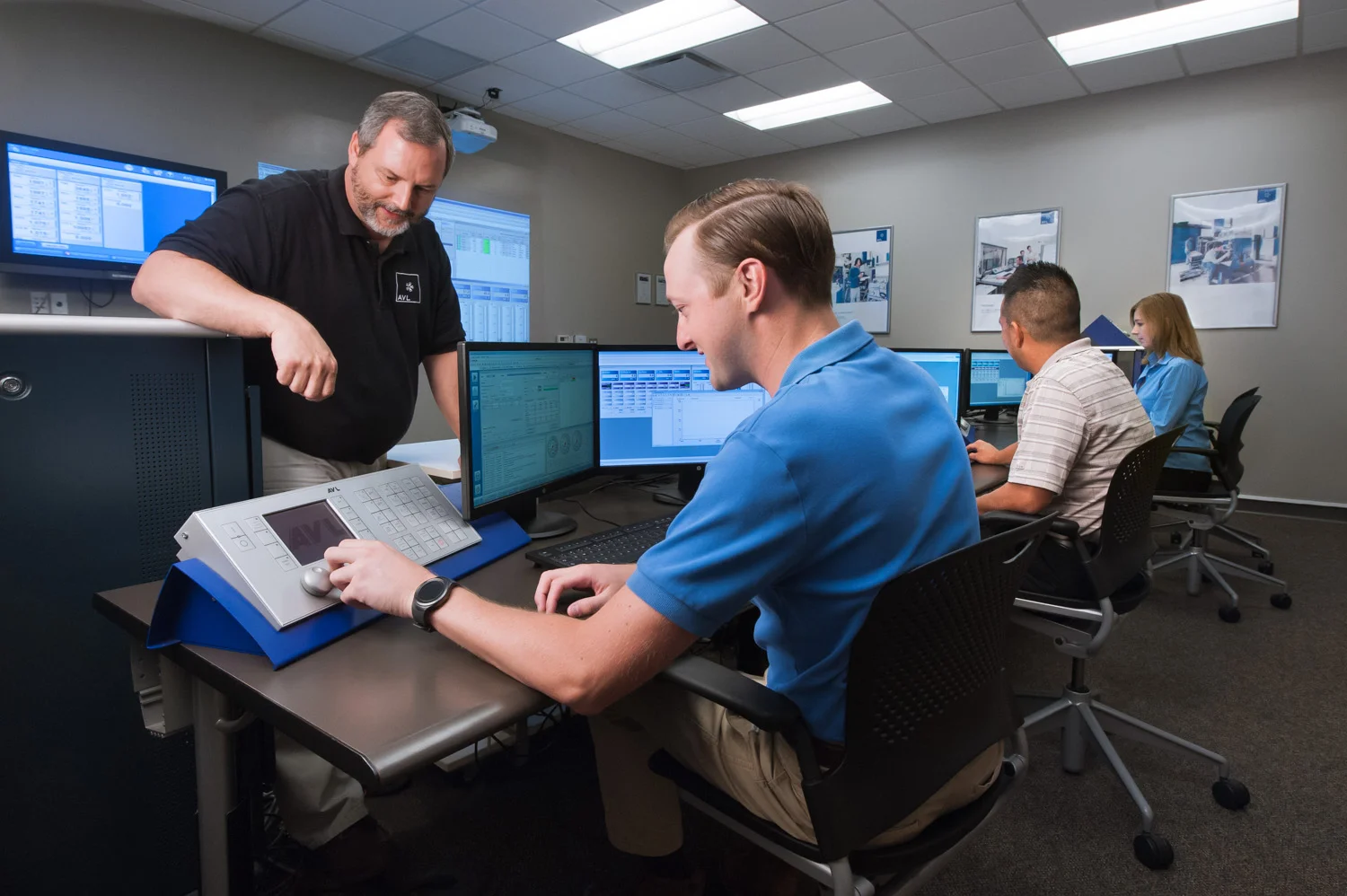 Office with four people working at desks with multiple computer monitors; one standing man shows something to three seated individuals.