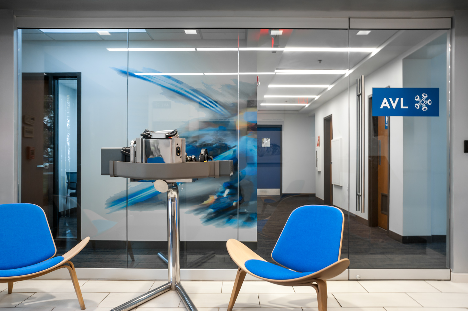 A waiting area with two blue chairs and a robotic device in front of a glass wall with an AVL sign, inside a modern office or facility.