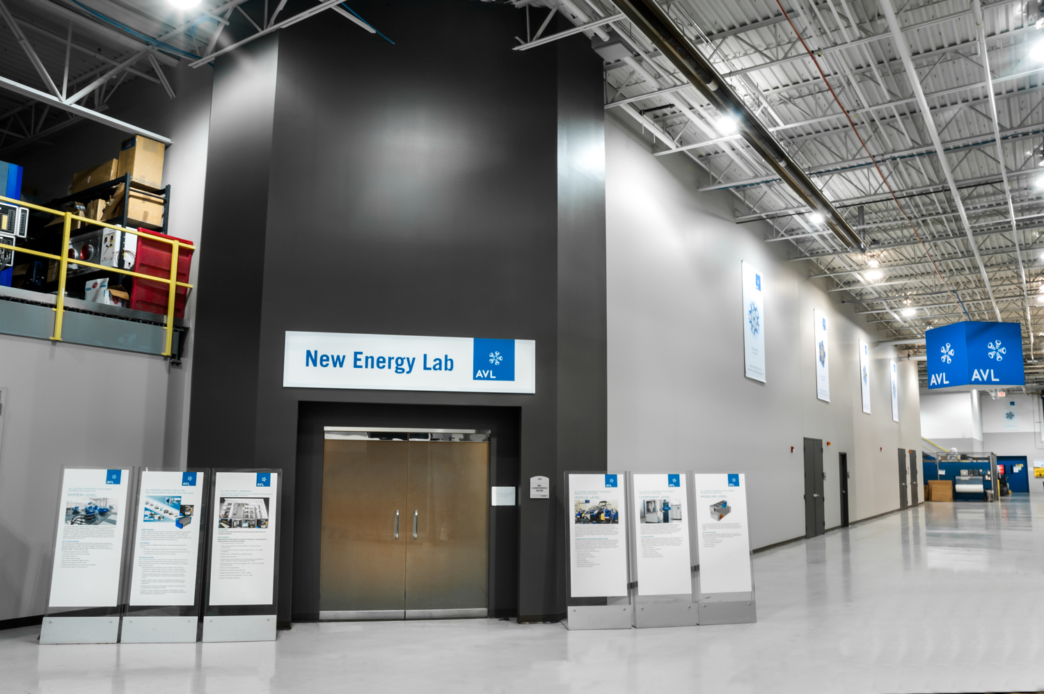 Entrance to the New Energy Lab at AVL, inside a large industrial facility with informational posters on easels and industrial equipment stored on a yellow-rimmed shelf.