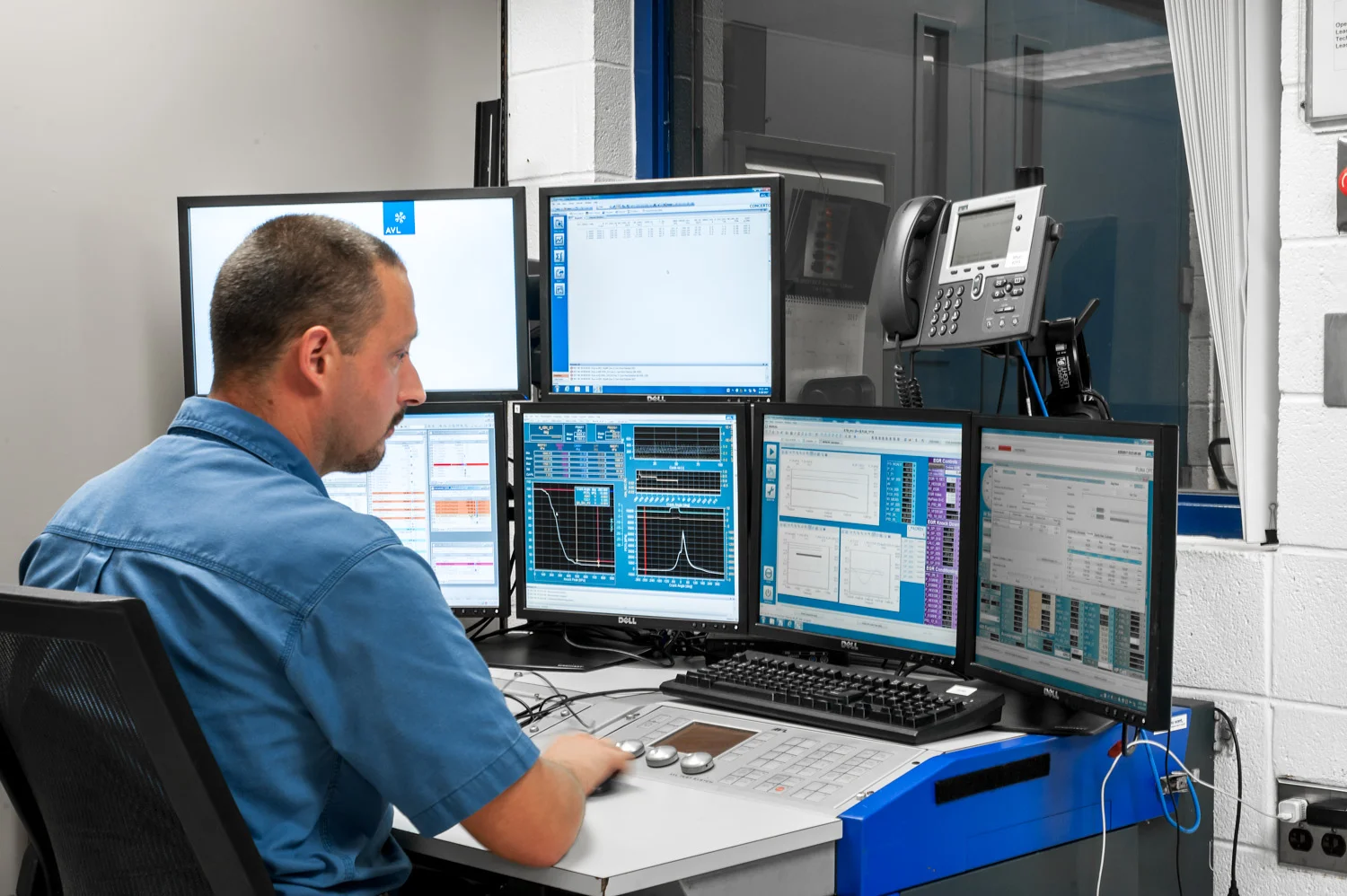 A man working at a desk with multiple computer monitors displaying technical data, graphs, and charts, in an industrial or office setting.