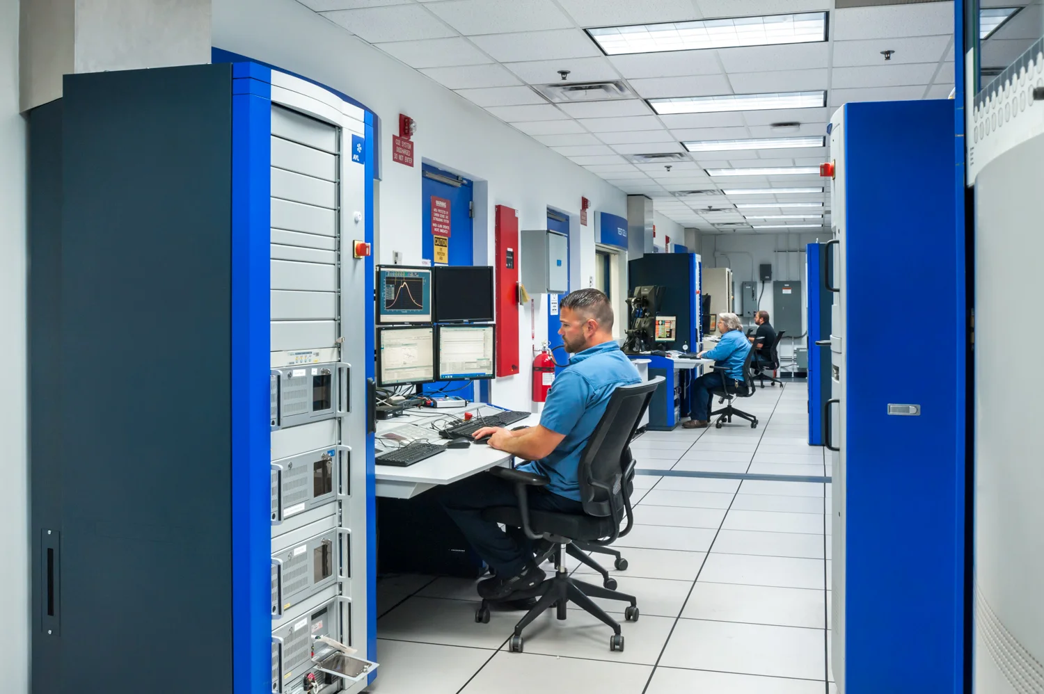 IT professionals working on multiple computer monitors in a high-tech control room with equipment racks and warning signs.