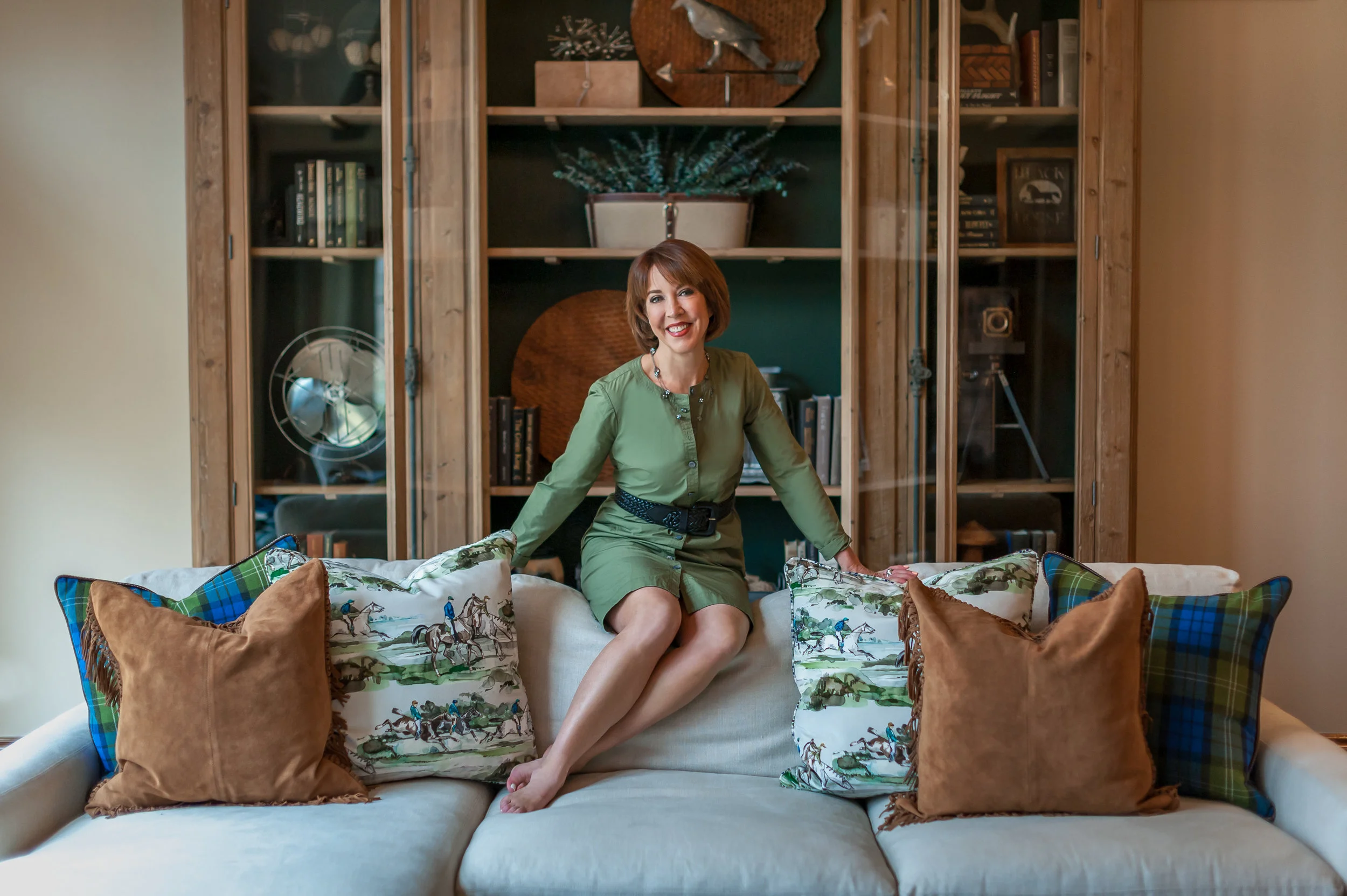A woman in a green dress sitting on a cream sofa with colorful throw pillows in a living room.