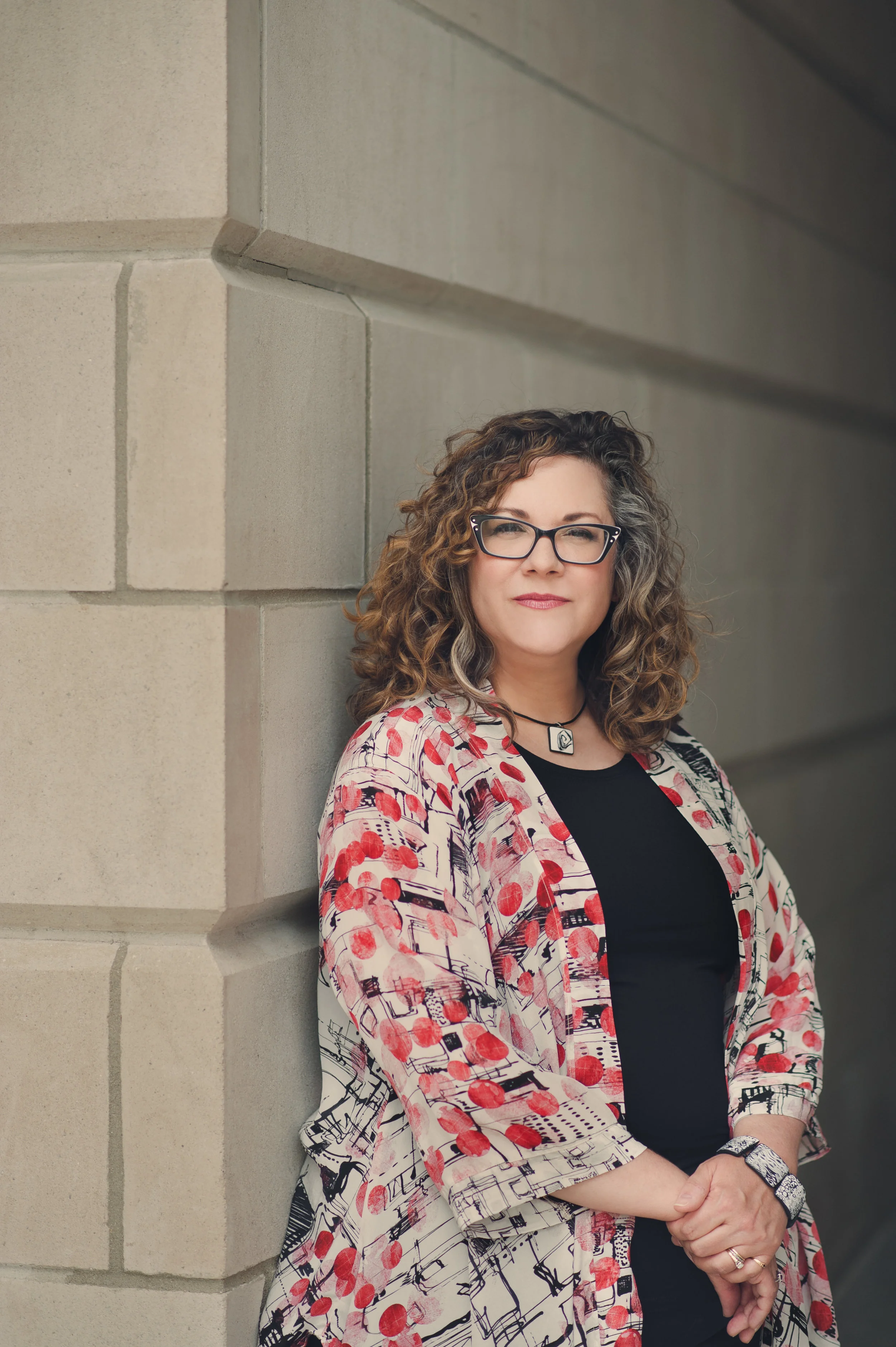 A woman with curly hair and glasses leaning against a stone wall, wearing a patterned jacket and black top.