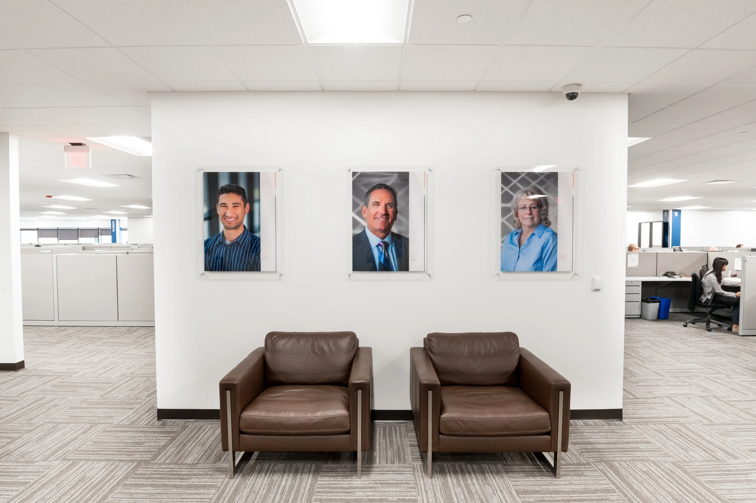 Office lobby with three framed portraits of diverse professionals on a white wall, brown leather chairs below, and open workspace with employees in the background.