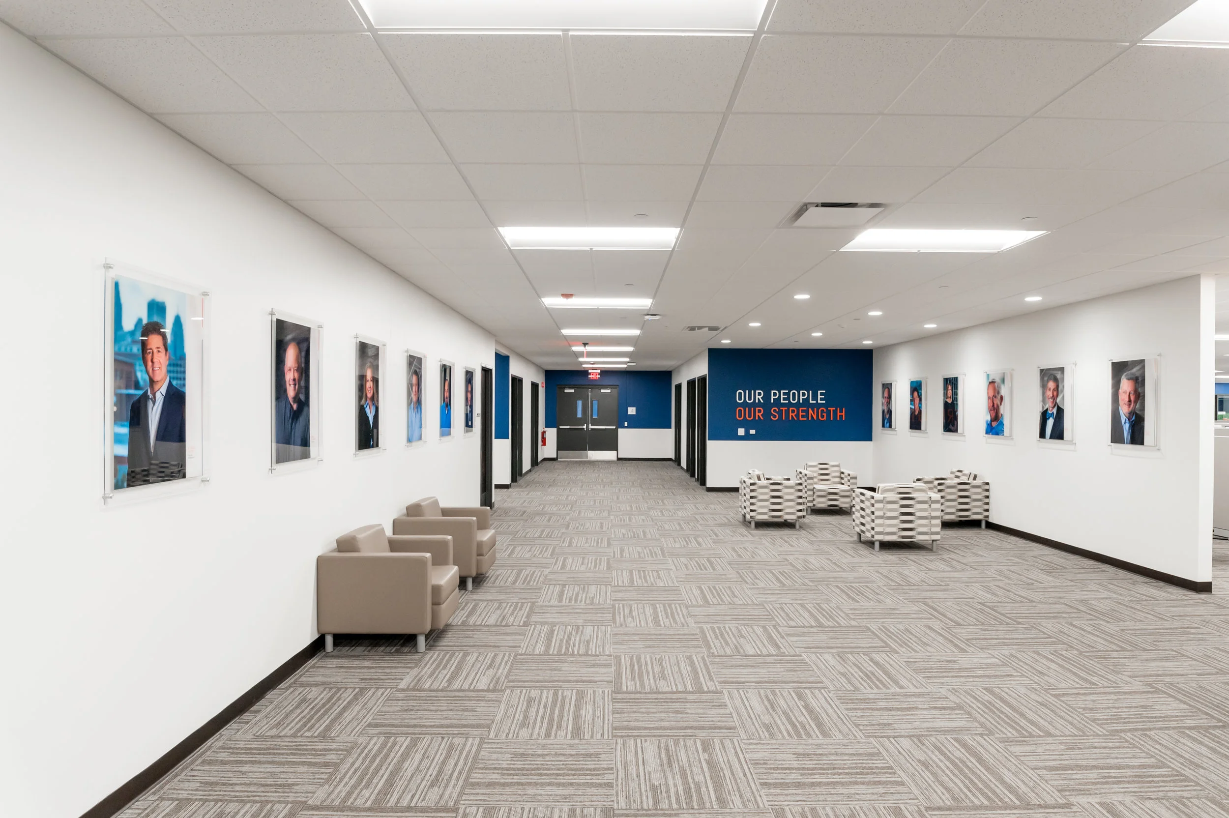 Lobby area with framed portraits of people on white walls, a seating area with beige and plaid armchairs, a blue accent wall with the sign 'Our People, Our Strength'.