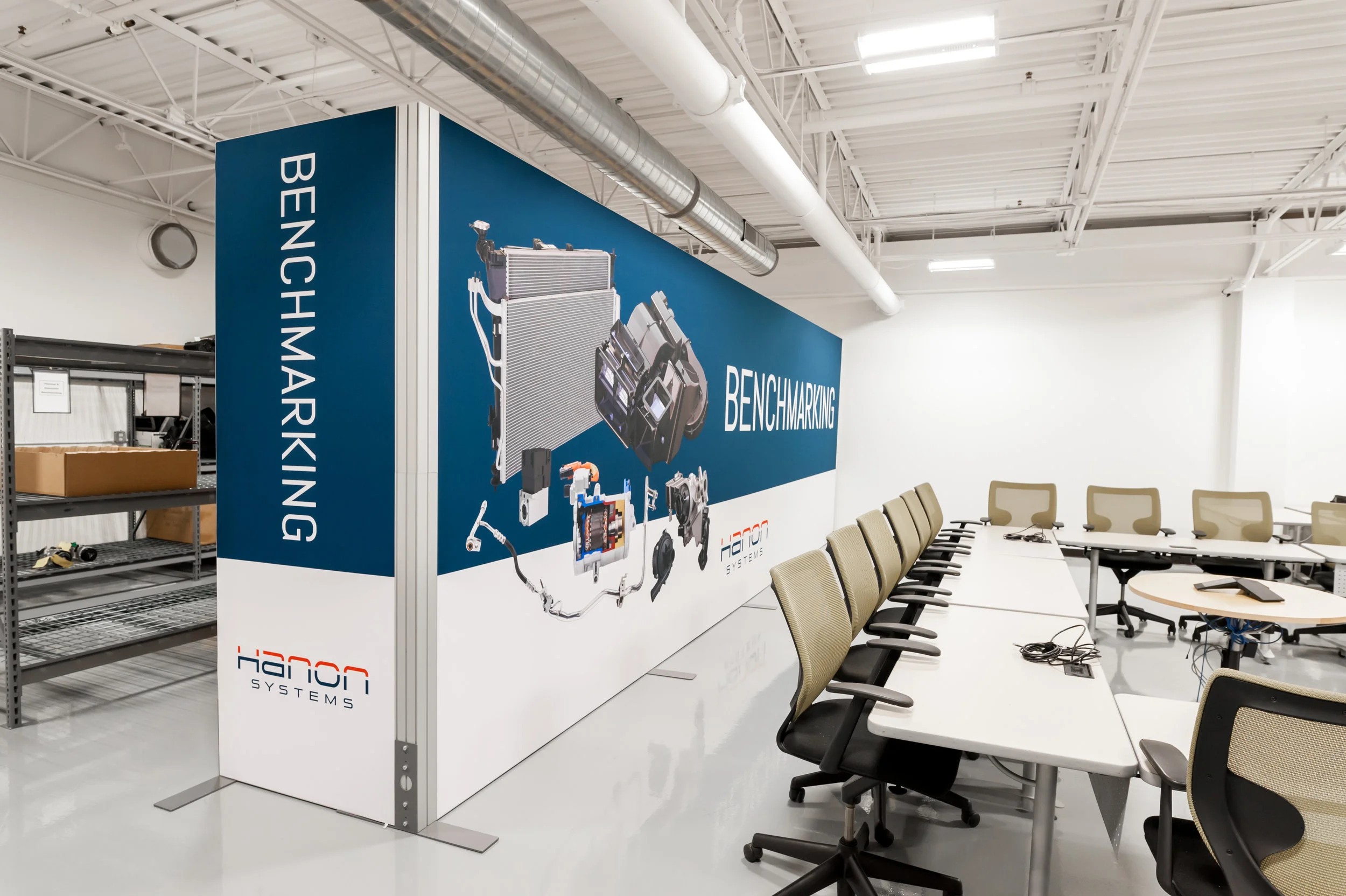 Empty conference room with beige chairs and white tables, and a large informational display about benchmarking at Hanon Systems.