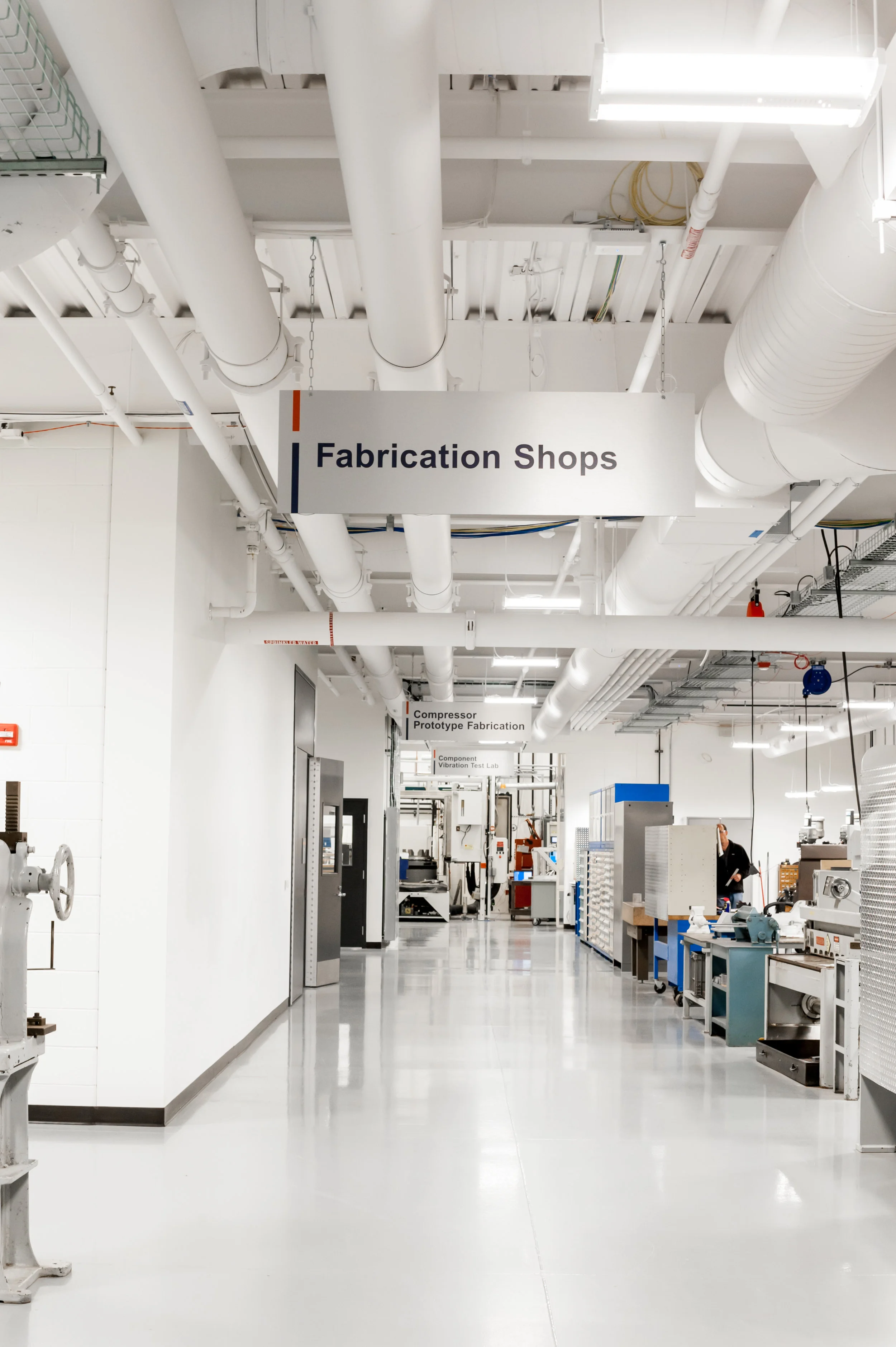 Interior of an industrial facility with signs for fabrication shops, including ductwork, machinery, and a person in the background.