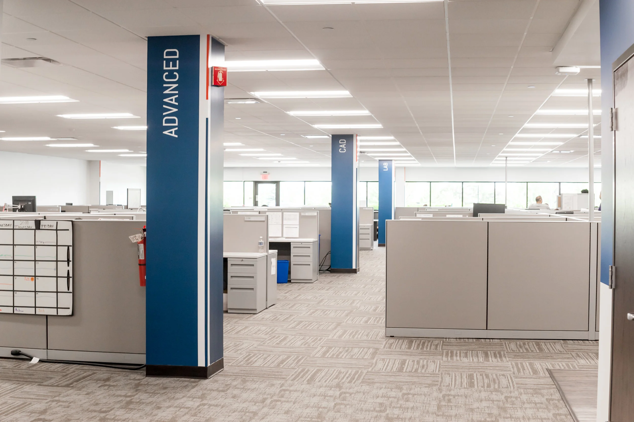 Empty modern office cubicles with blue and white signs labeled 'ADVANCED' and 'CAD', and a whiteboard with a weekly schedule on the left.