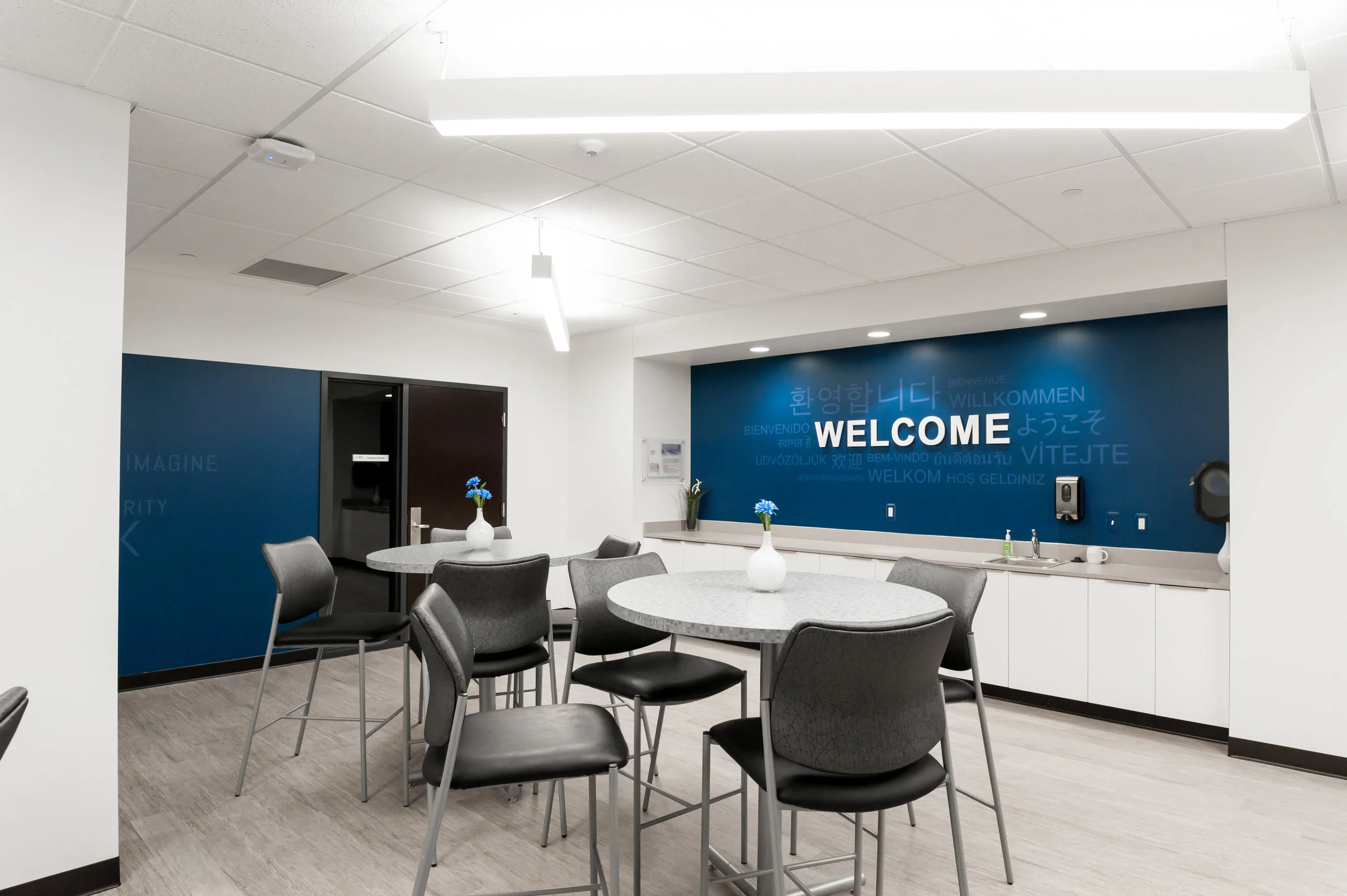 An indoor common area with round tables, gray chairs, blue vases with flowers, a blue accent wall with 'WELCOME' and greeting words in multiple languages, and a kitchenette with a sink, soap dispenser, and cups.