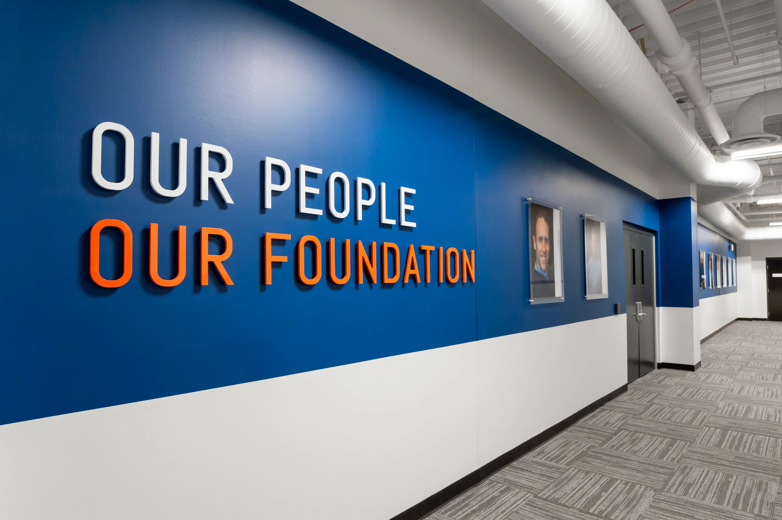 Hallway with blue and white walls, a sign that says "Our People Our Foundation" in white and orange 3D letters, framed portraits on the wall, and a closed metal door.