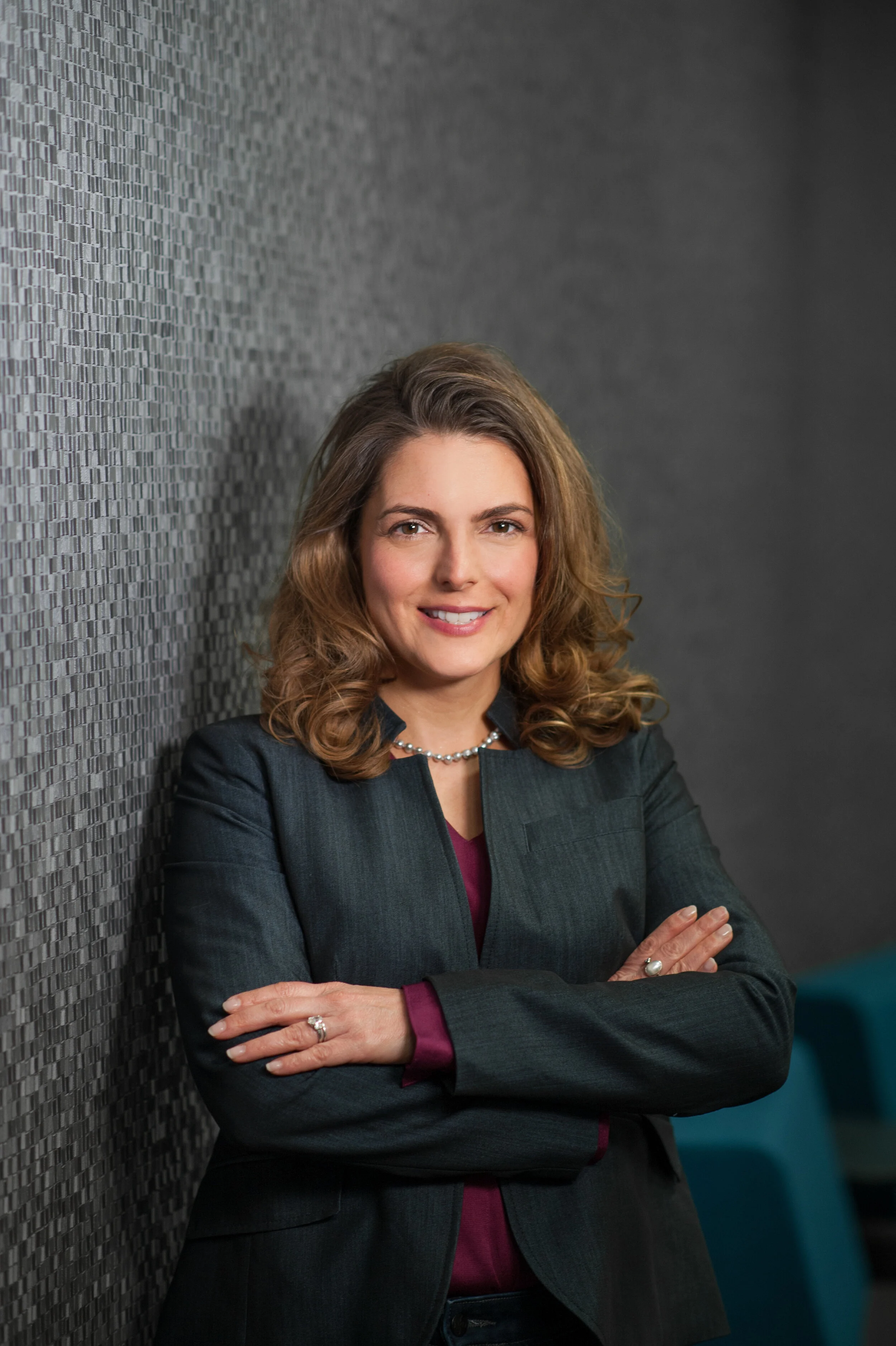 Portrait of a woman with wavy brown hair wearing a dark blazer and a pearl necklace, standing with arms crossed against a textured gray wall.