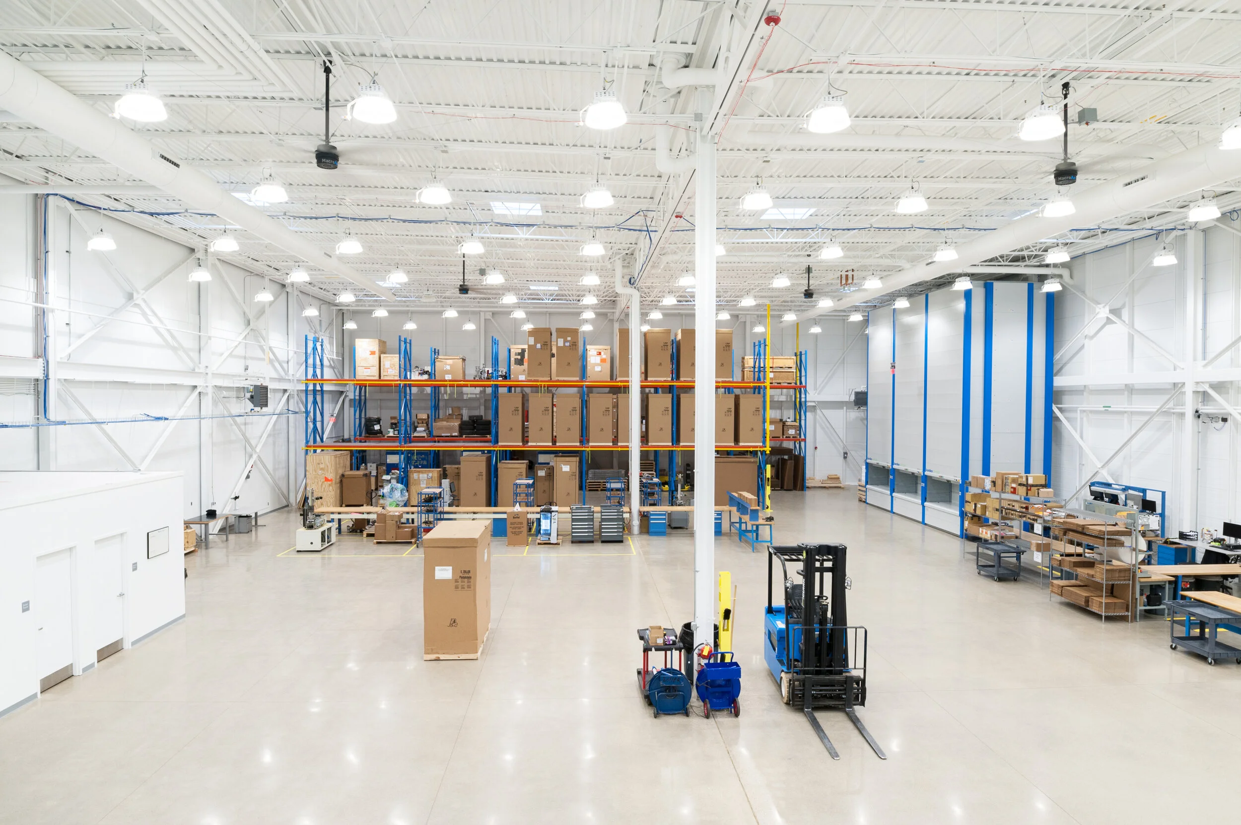 Interior of a spacious warehouse with high ceilings, bright lighting, and organized shelves filled with boxes and equipment, along with a forklift in the foreground.
