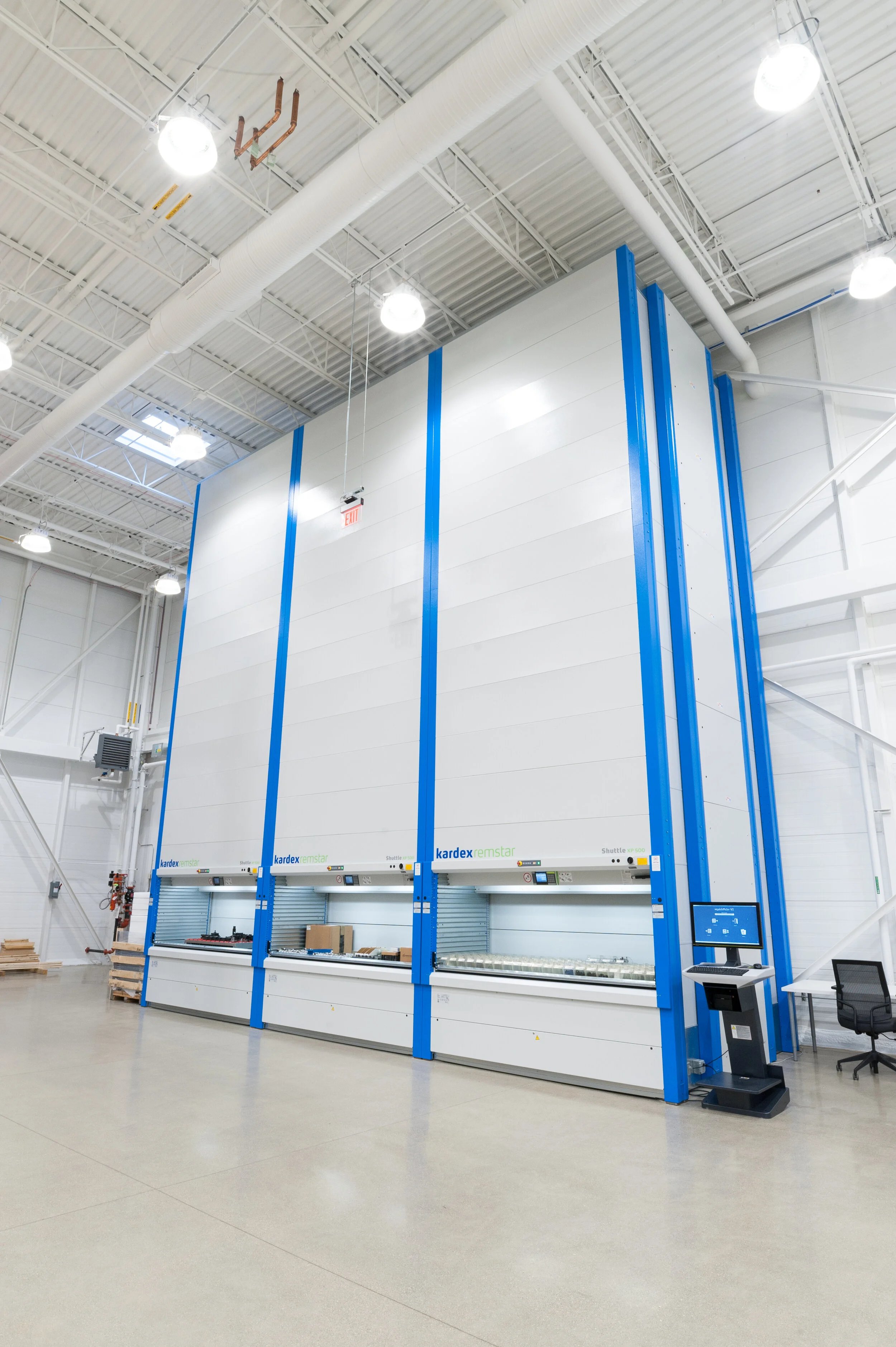 A large industrial warehouse with high ceiling, bright lighting, and an automated storage and retrieval system with blue and white structural elements.