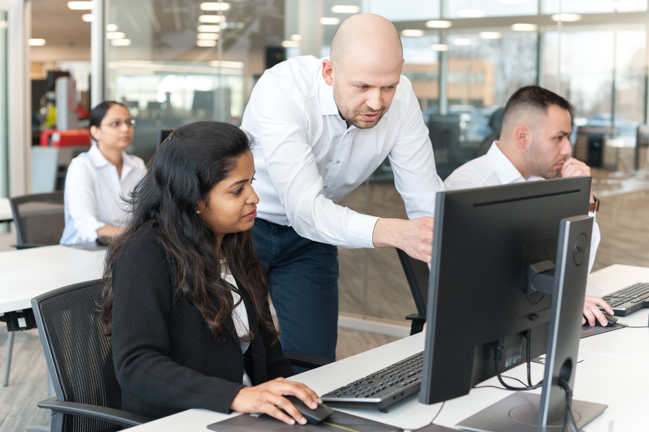 A male coworker helps a female employee with her computer in an office setting while two other employees work at their desks in the background.
