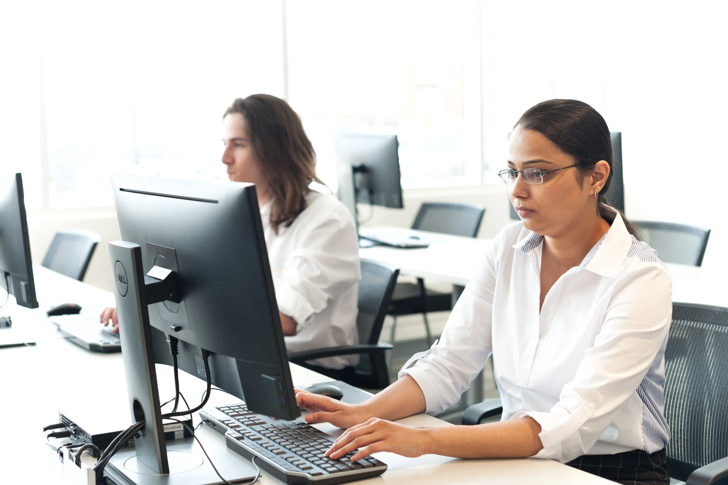 Two women working on desktop computers in an office with large windows and bright natural light