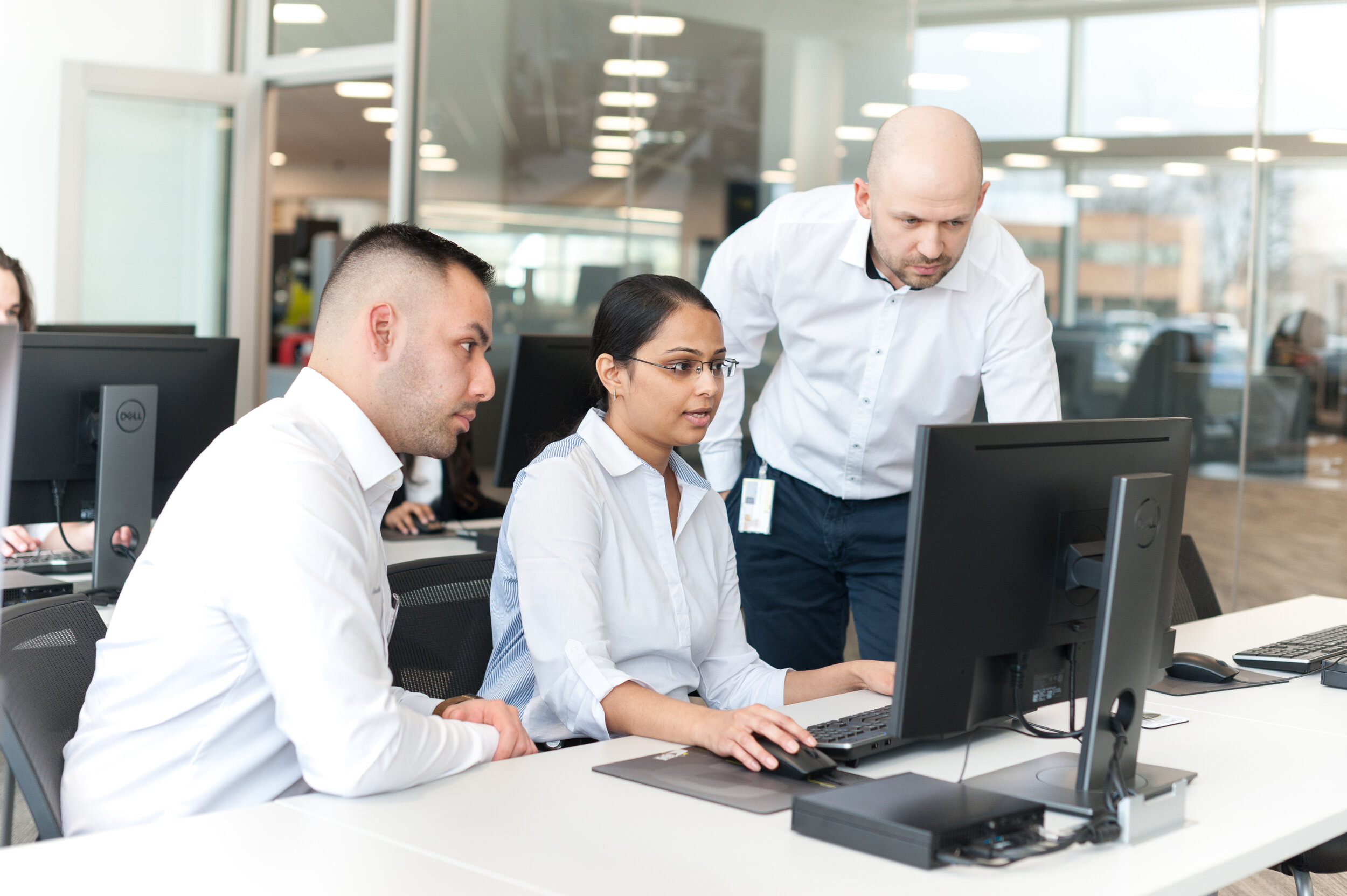 Office scene with three employees working together at a desk with computers, in a well-lit modern office environment.