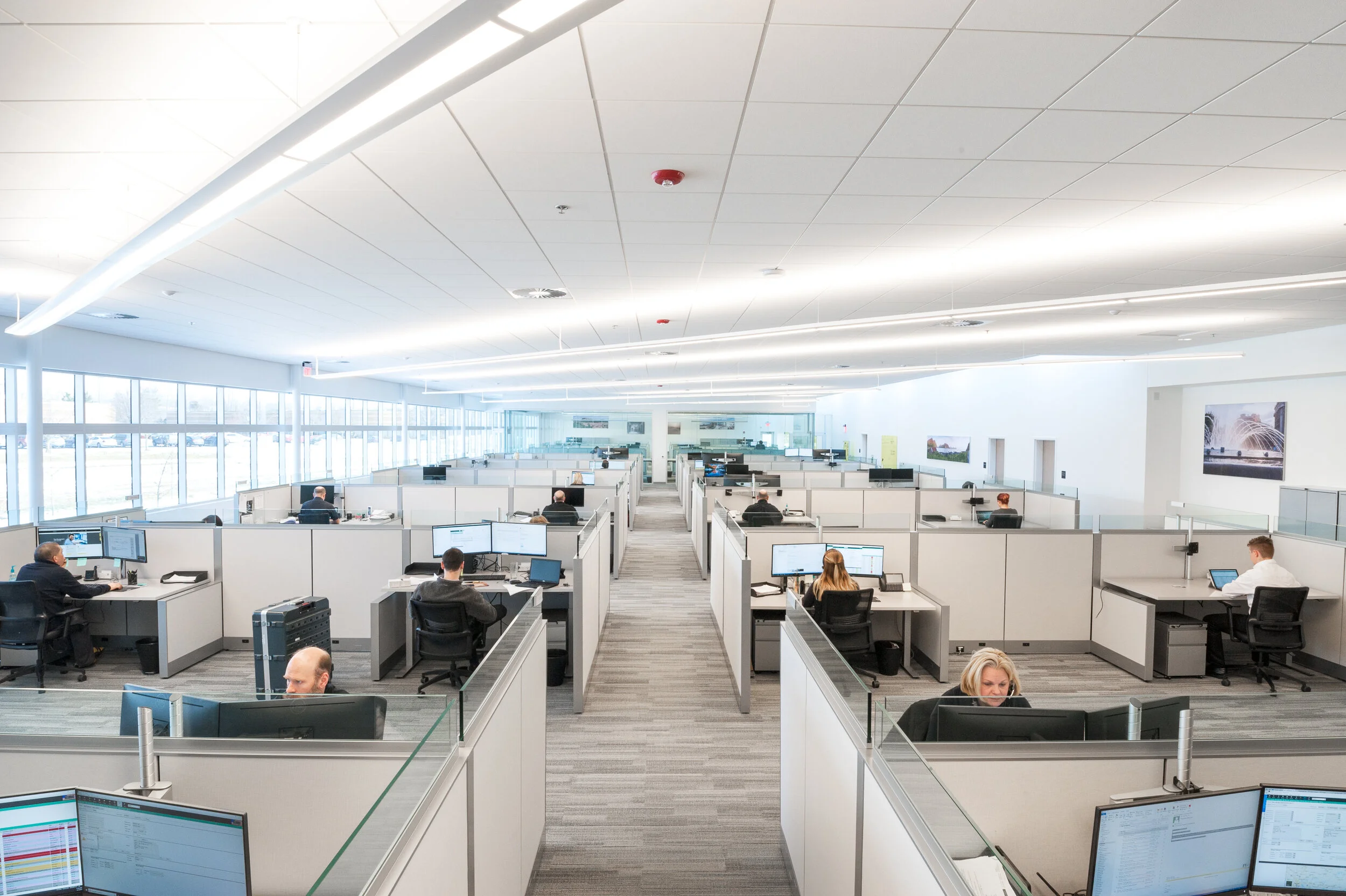 Open-plan office with multiple employees working at cubicles with computers and monitors, large windows along the side, modern lighting, and a bright, spacious environment.
