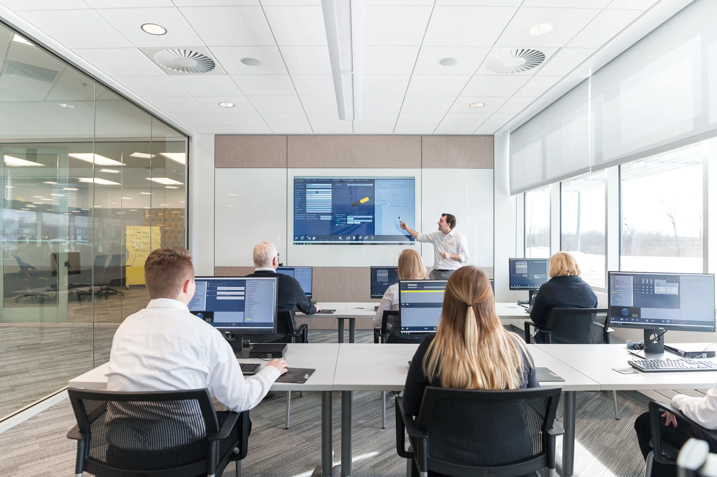 A business trainer or presenter instructing a group of adults in a modern, well-lit conference room using a digital whiteboard connected to computers.