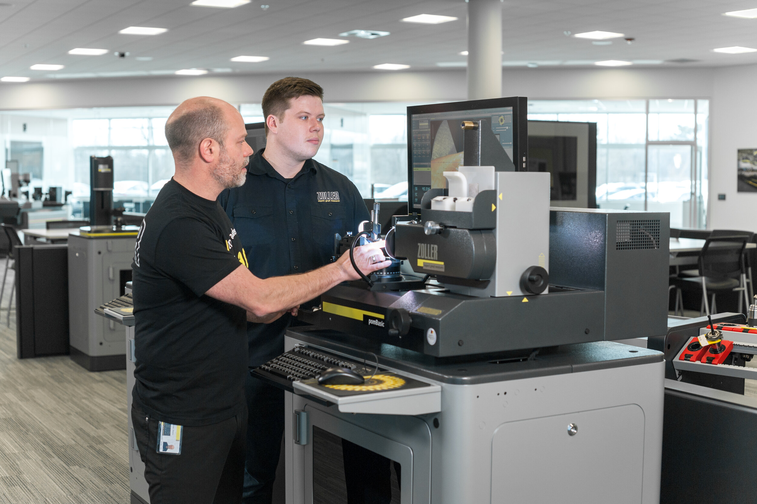 Two men working with a laser testing machine in an office or showroom. One man is operating the machine while the other observes. There are monitors and other testing equipment visible.