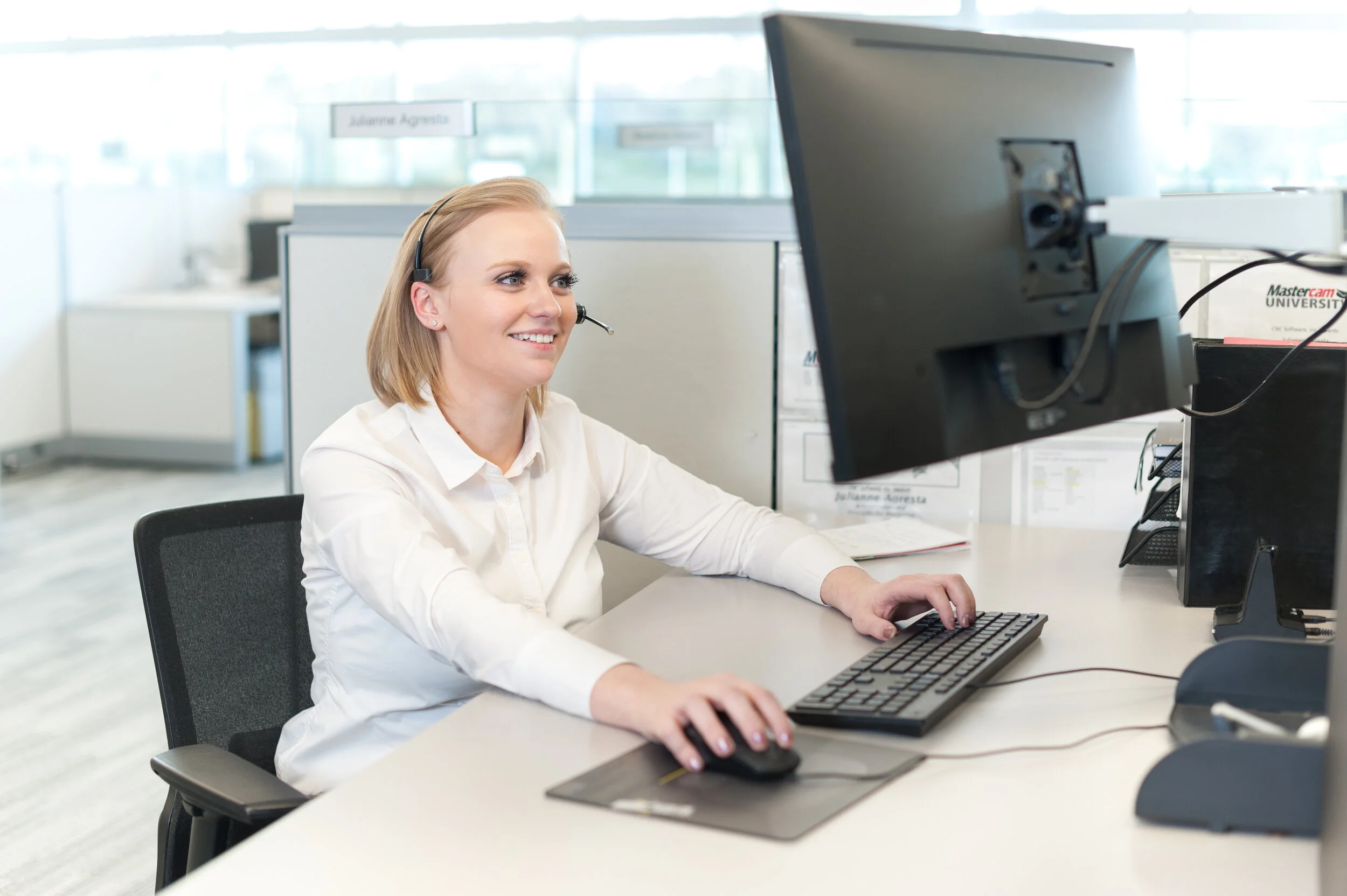 A woman with blonde hair wearing a white shirt and headset working at a computer in an office.