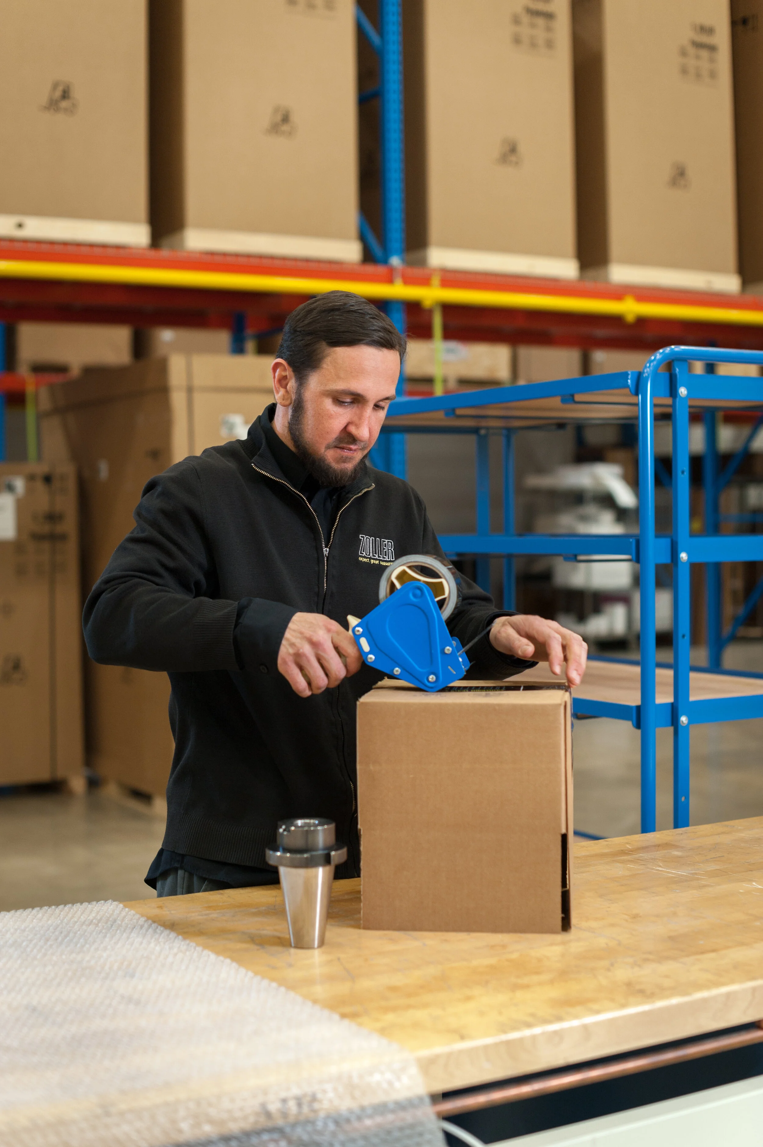 A man packing a cardboard box using packing tape in a warehouse with shelves of large boxes in the background.