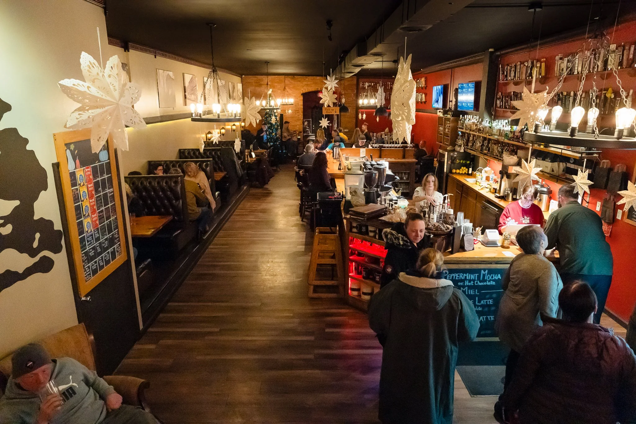 A cozy cafe with customers sitting at tables and the bar, decorated with large paper snowflakes hanging from the ceiling, with warm lighting and holiday decorations.