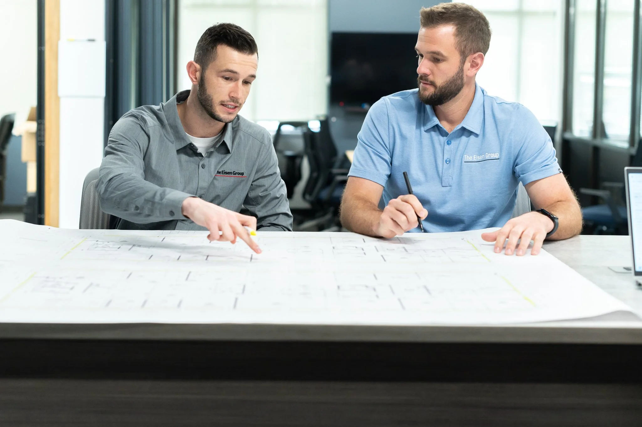 Two men sitting at a table reviewing a blueprint or architectural plan in an office setting.