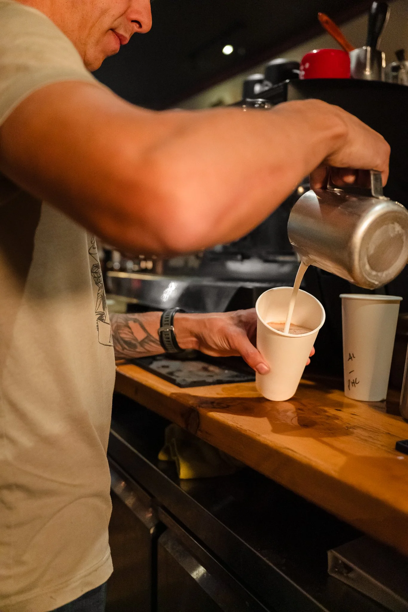A person pouring coffee or tea from a metal pitcher into a white paper cup at a café or coffee shop.