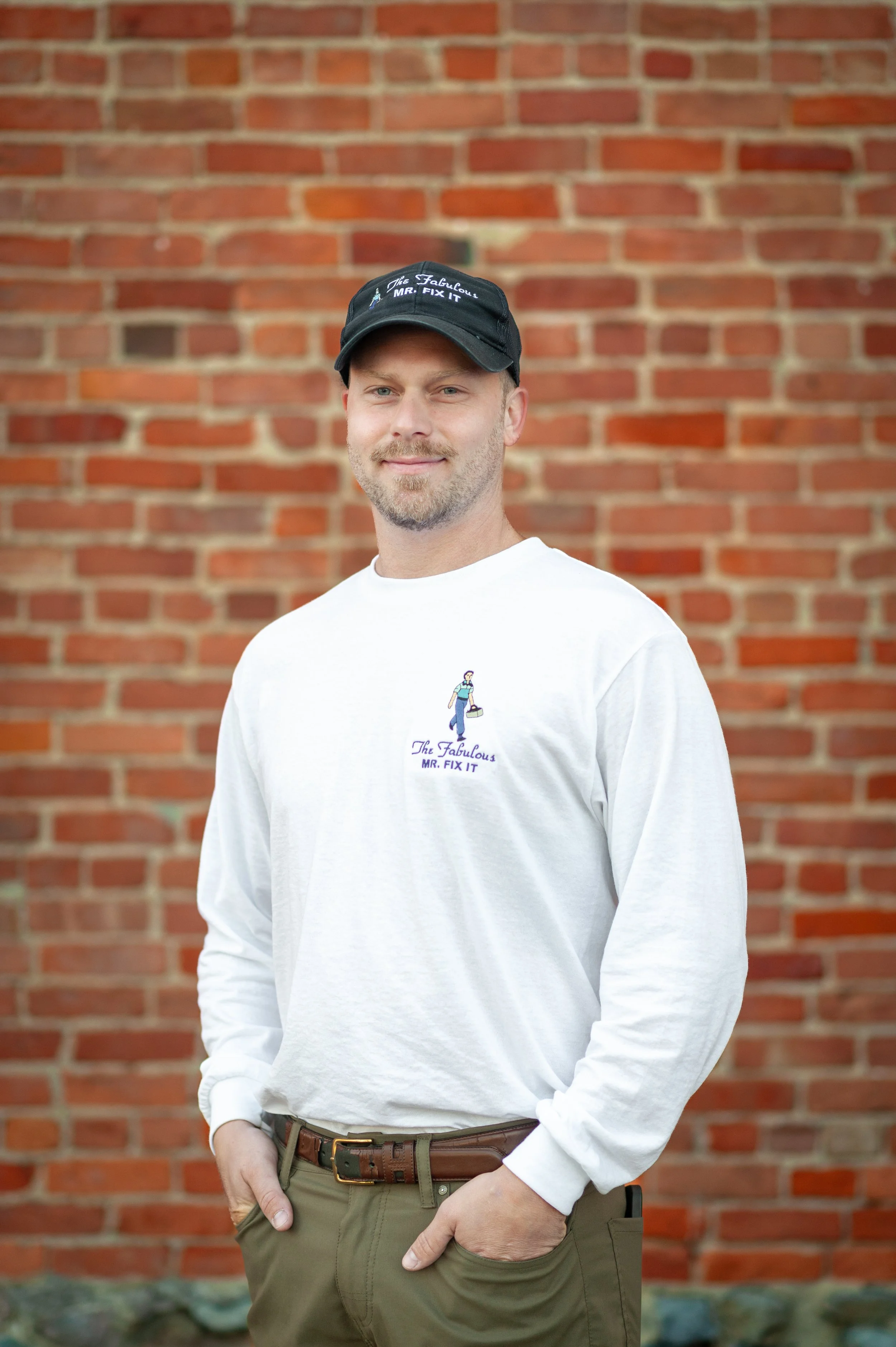 A man standing outdoors in front of a brick wall. He is wearing a black cap and a white long-sleeved shirt with a logo that reads 'The Fabulous Mr. Fix It' and features a cartoon illustration of a person holding a wrench and a toolbox. He has a beard