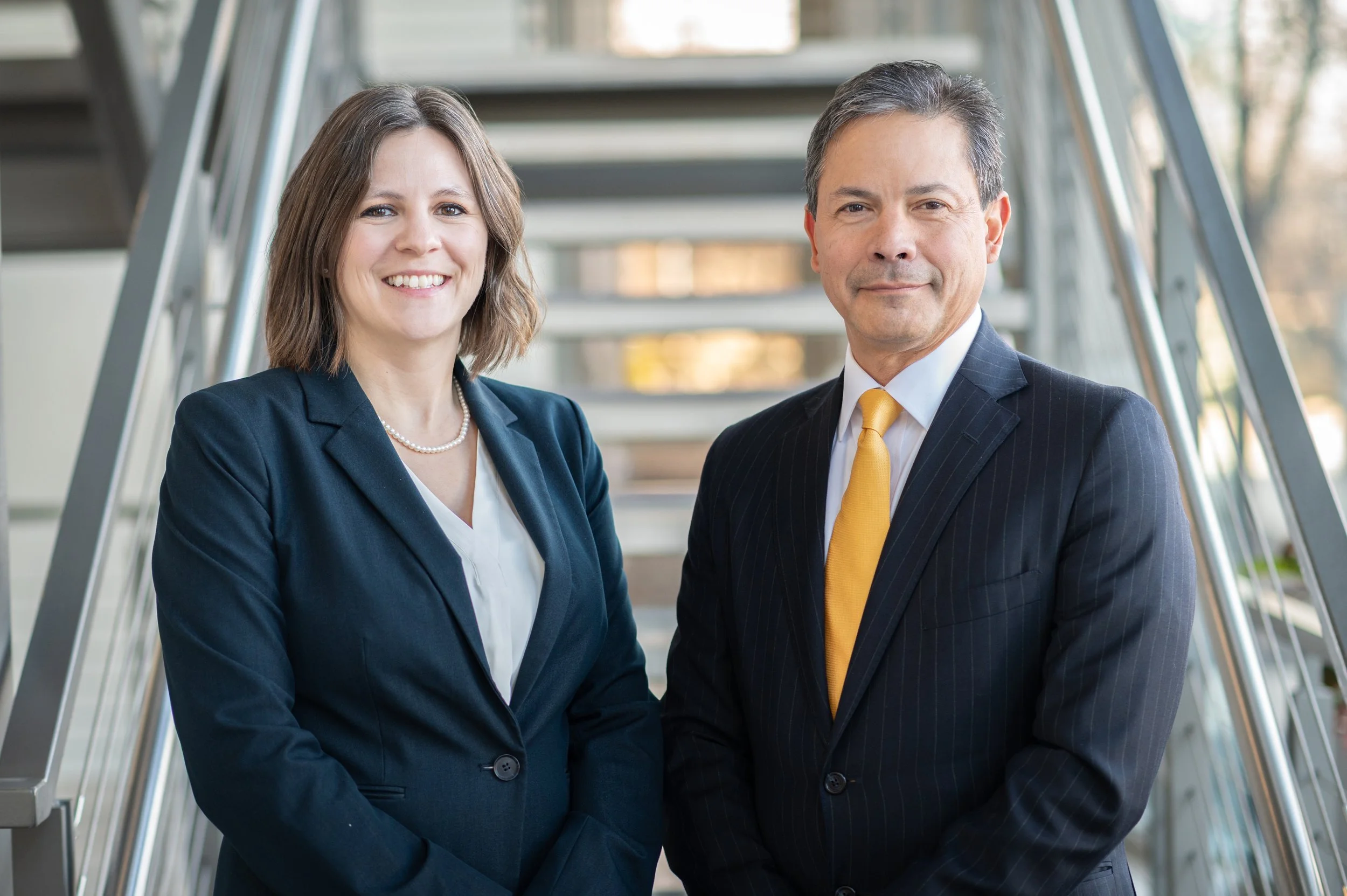 Two professionally dressed individuals, a woman and a man, standing side by side on a staircase in an office building, smiling at the camera.
