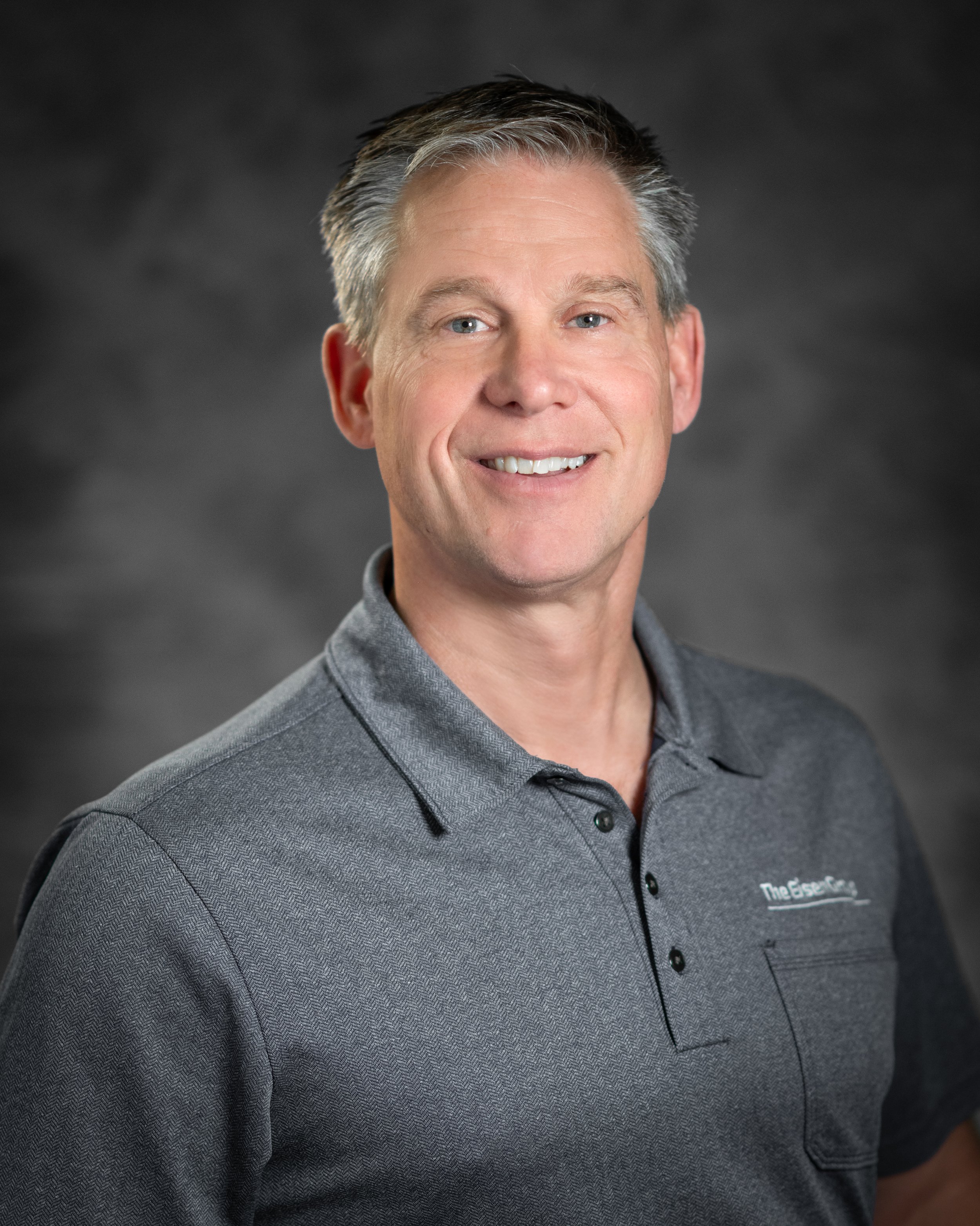 Professional portrait of a smiling middle-aged man with gray hair, wearing a gray polo shirt, against a dark gray background.