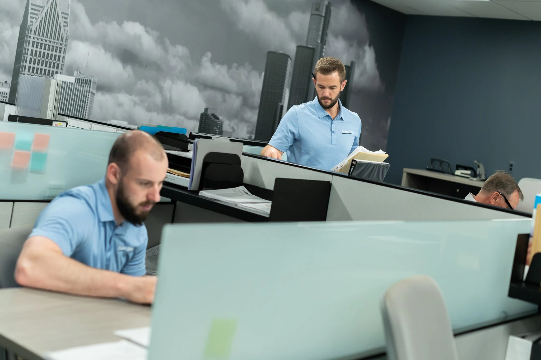Three men working at cubicles in an office with a city skyline mural featuring skyscrapers and clouds on the wall.