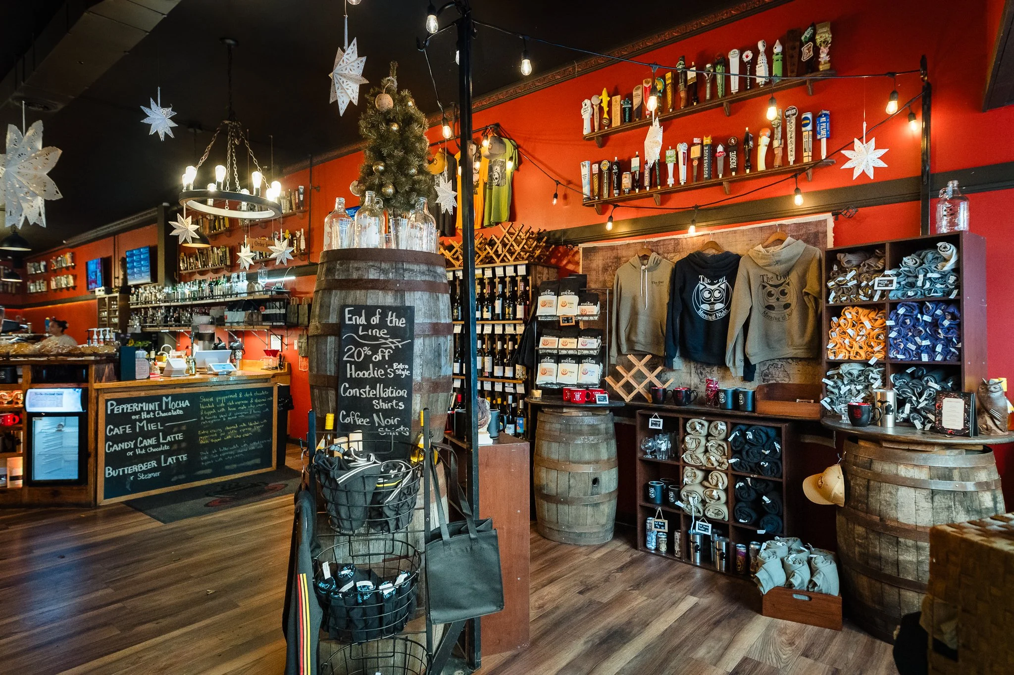 Interior of a cozy coffee shop decorated for Christmas with hanging snowflakes and a small Christmas tree. Display shelves with wine bottles, hoodies, cones, and mugs are visible, along with a chalkboard sign offering discounts.