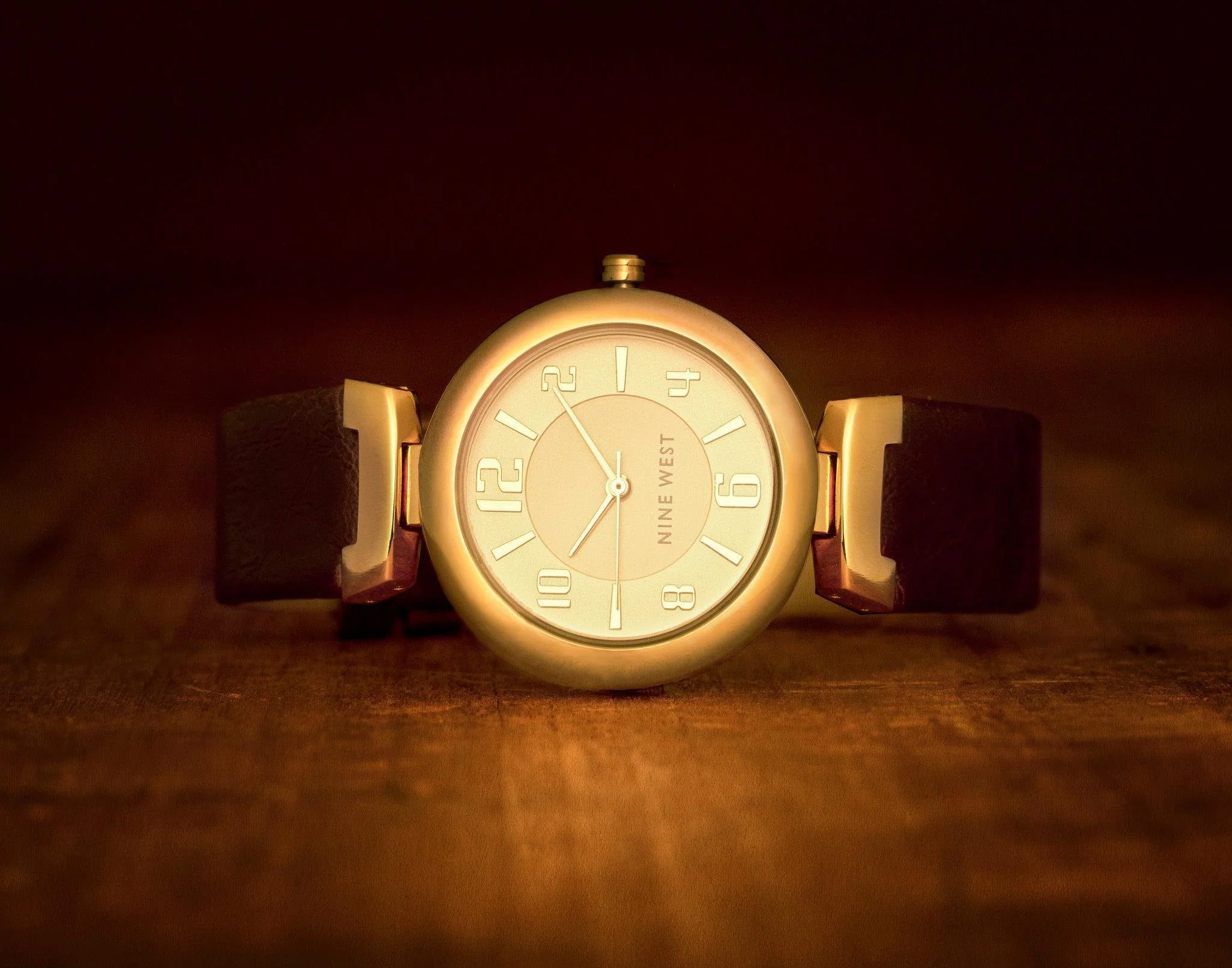 A wristwatch with a round face displaying the time, placed on a wooden surface with a dark background. The watch has a leather strap and a gold-colored bezel, with the brand name 'NINE WEST' on the dial.