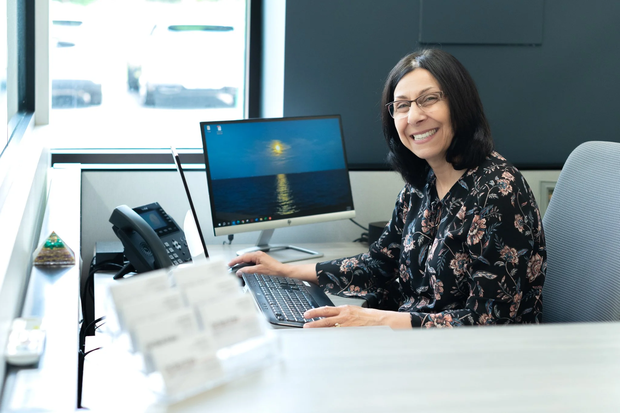 Smiling woman with glasses sitting at a desk in an office, working on a computer with a monitor displaying a sunset over the ocean.